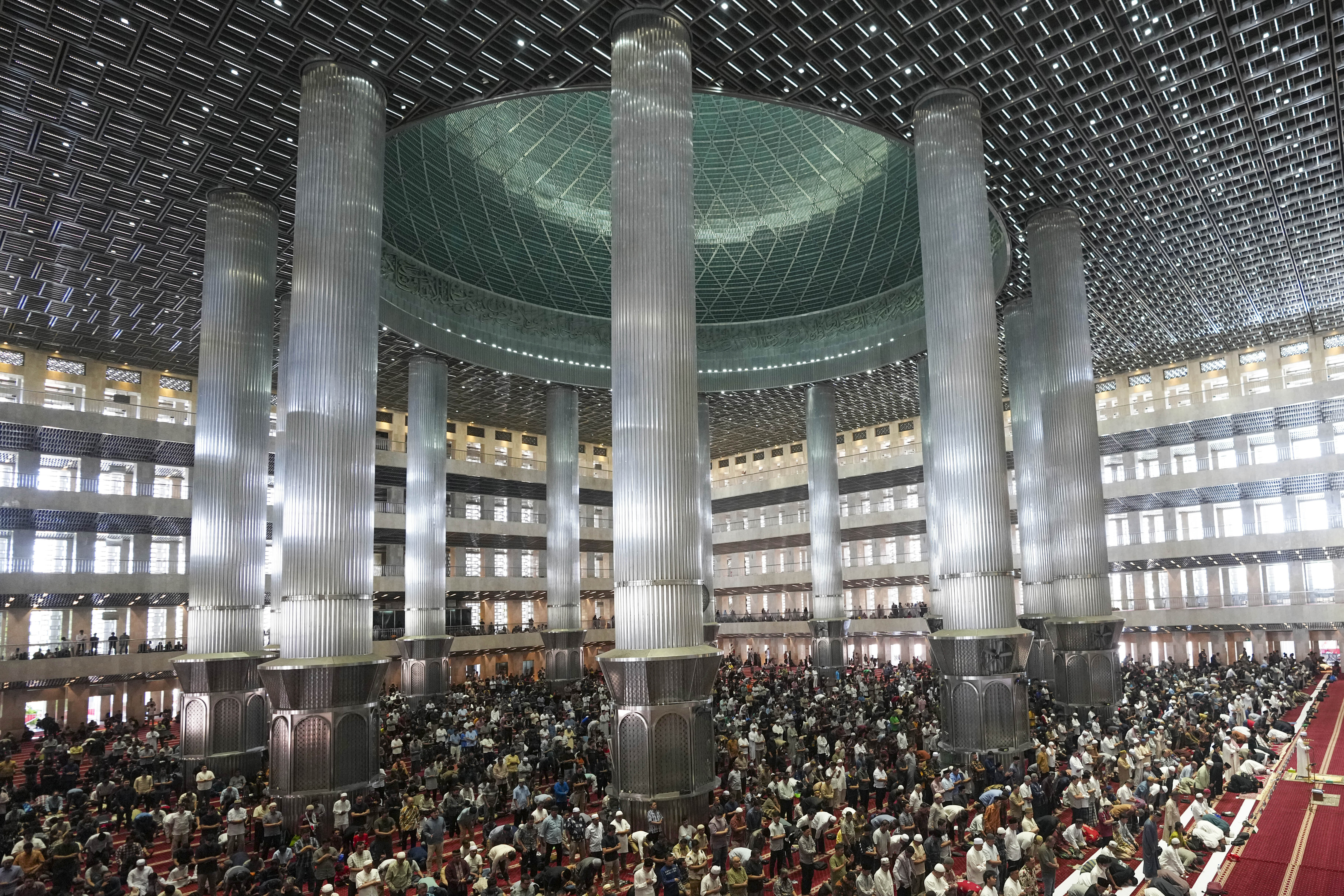 Muslims perform Friday prayer at Istiqlal Mosque in Jakarta. There are tall columns around the inside and a soaring roof. Light is falling onto the interior from the outside. 