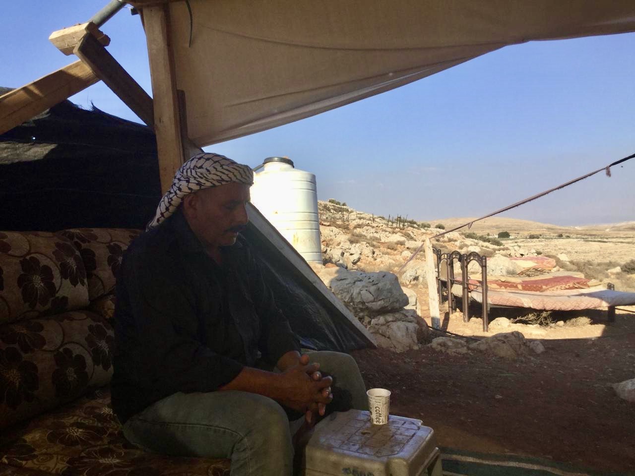 Abu Bashar sits outside under a canopy with a bed in the background