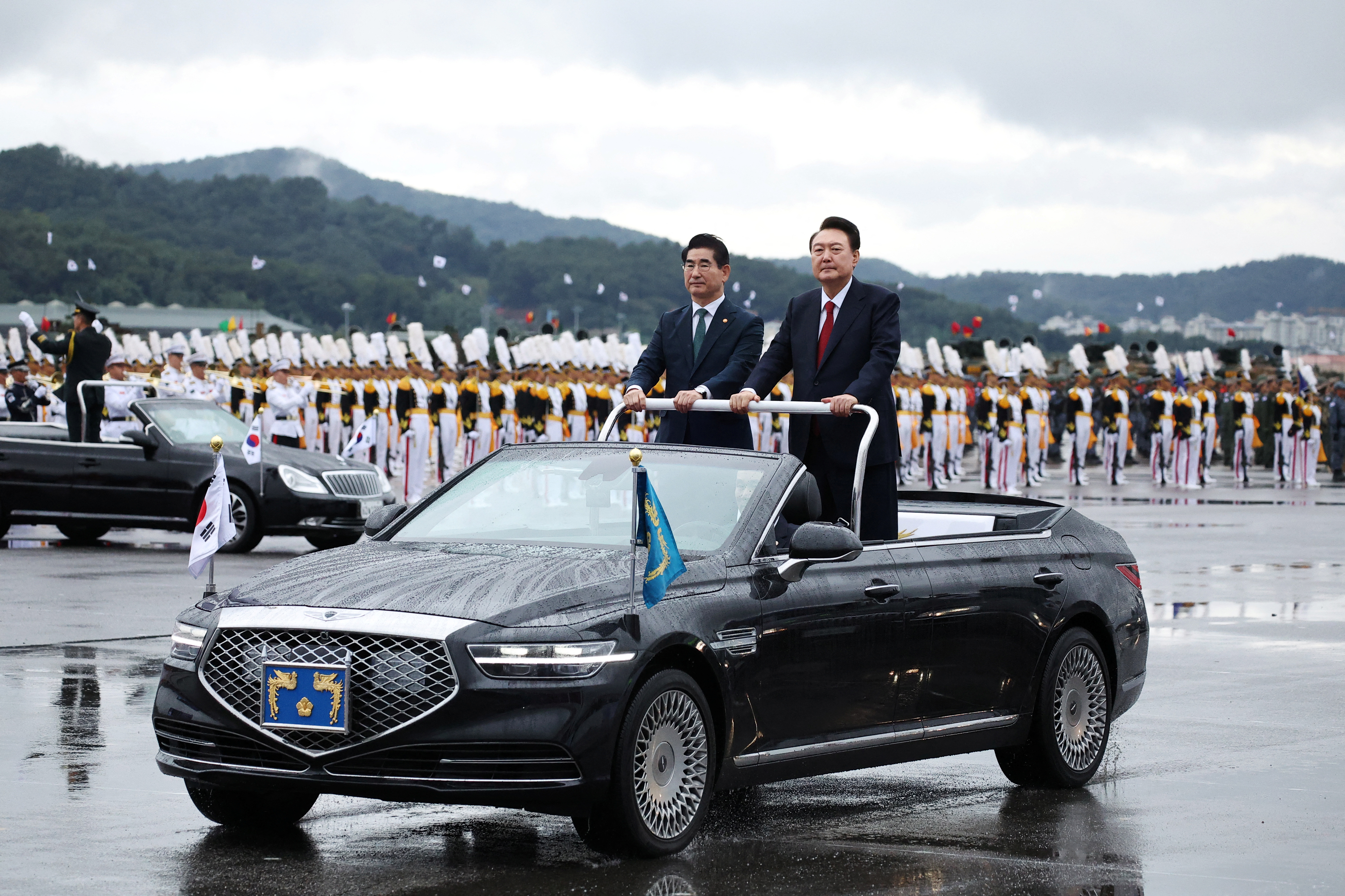 South Korean President Yoon Suk Yeol inspects South Korean troops in Seongnam. He is standing in an open topped car.with pennants on each side. The tarmac is wet from rain.