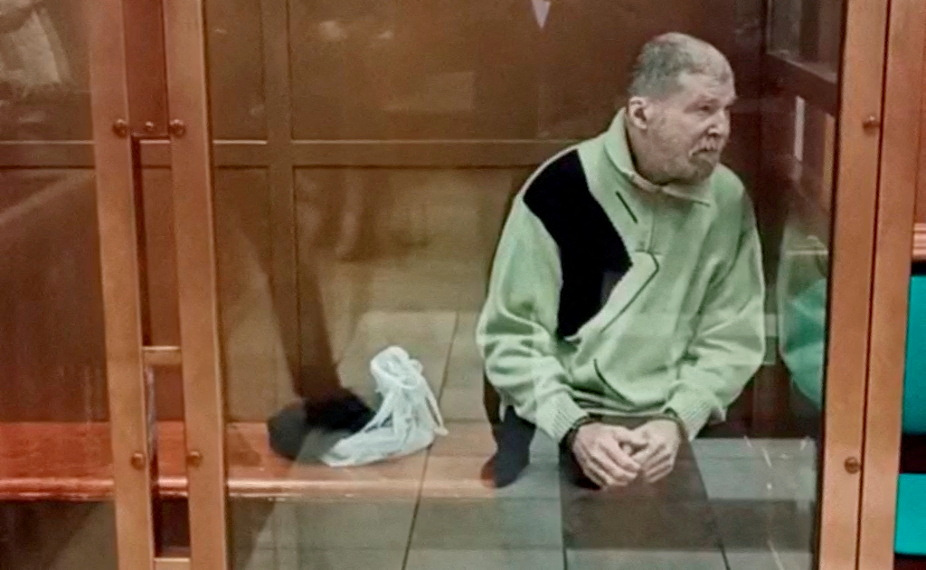 Stephen Hubbard in a glass walled dock in court. He is wearing a pale green top. His hands are cuffed and his head has been shaved.