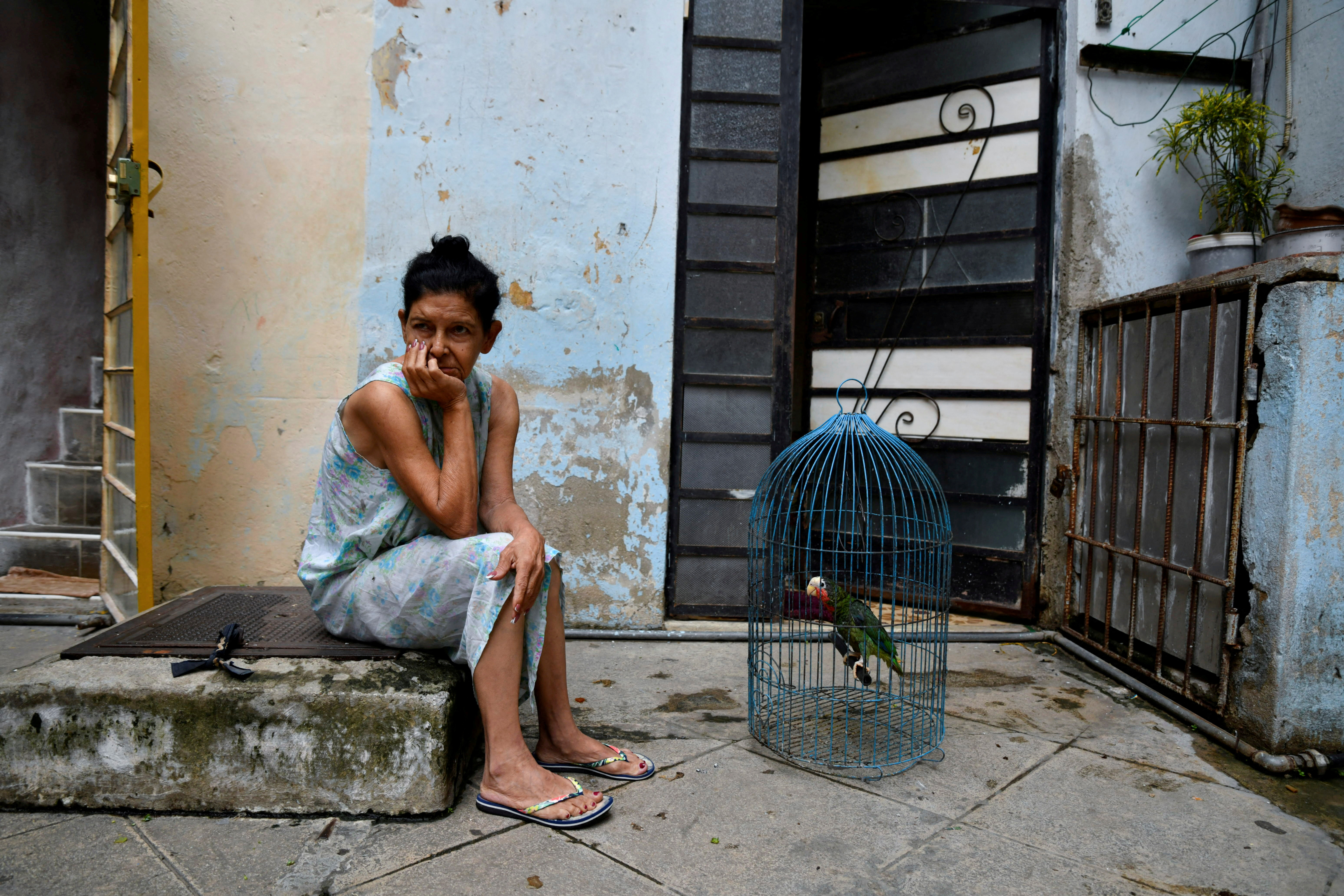 A woman sits on a stoop beside a bird cage with a parrot in Havana, her head in her hands.