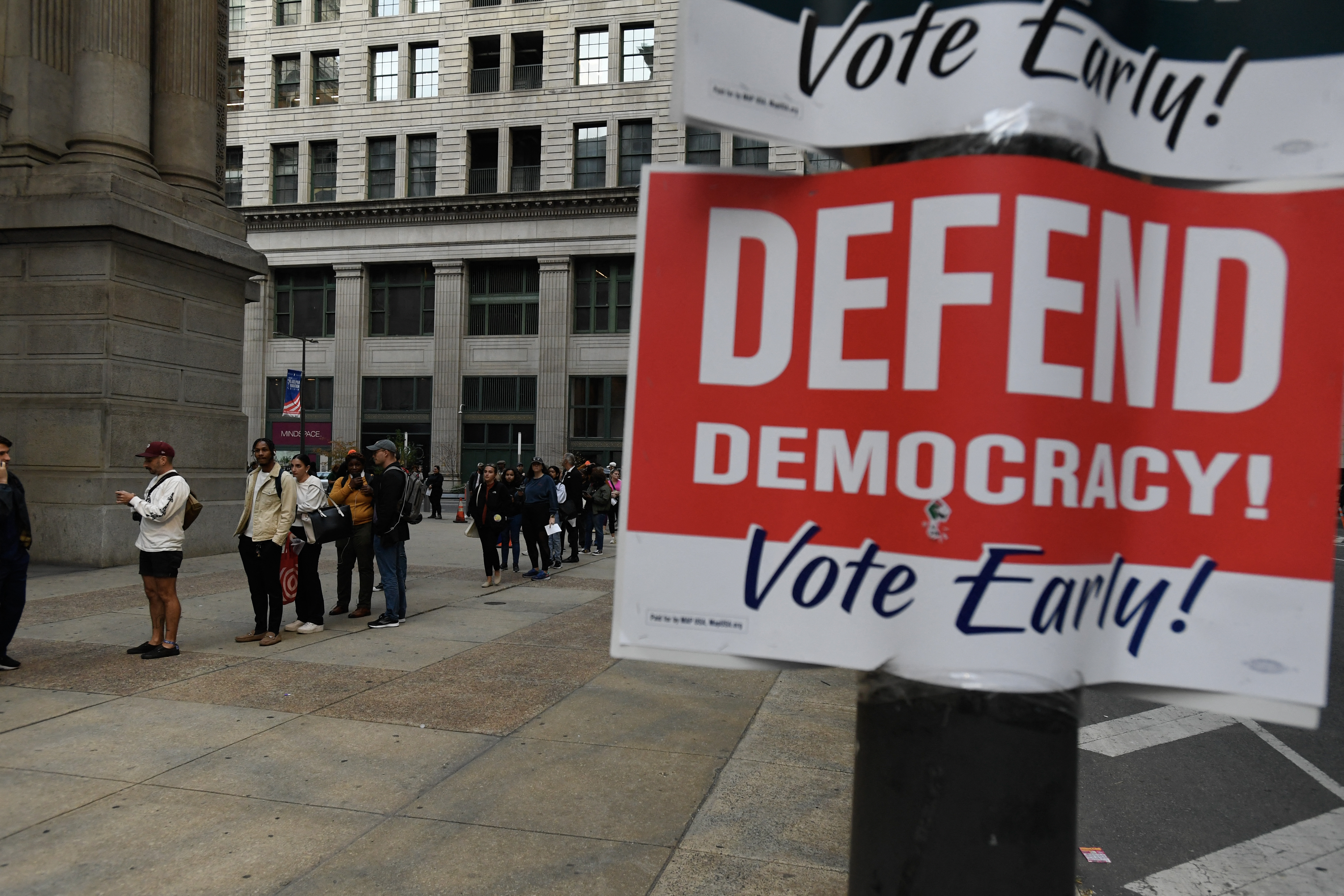 a red sign on a poll says defend democracy vote early as people line up in the distance