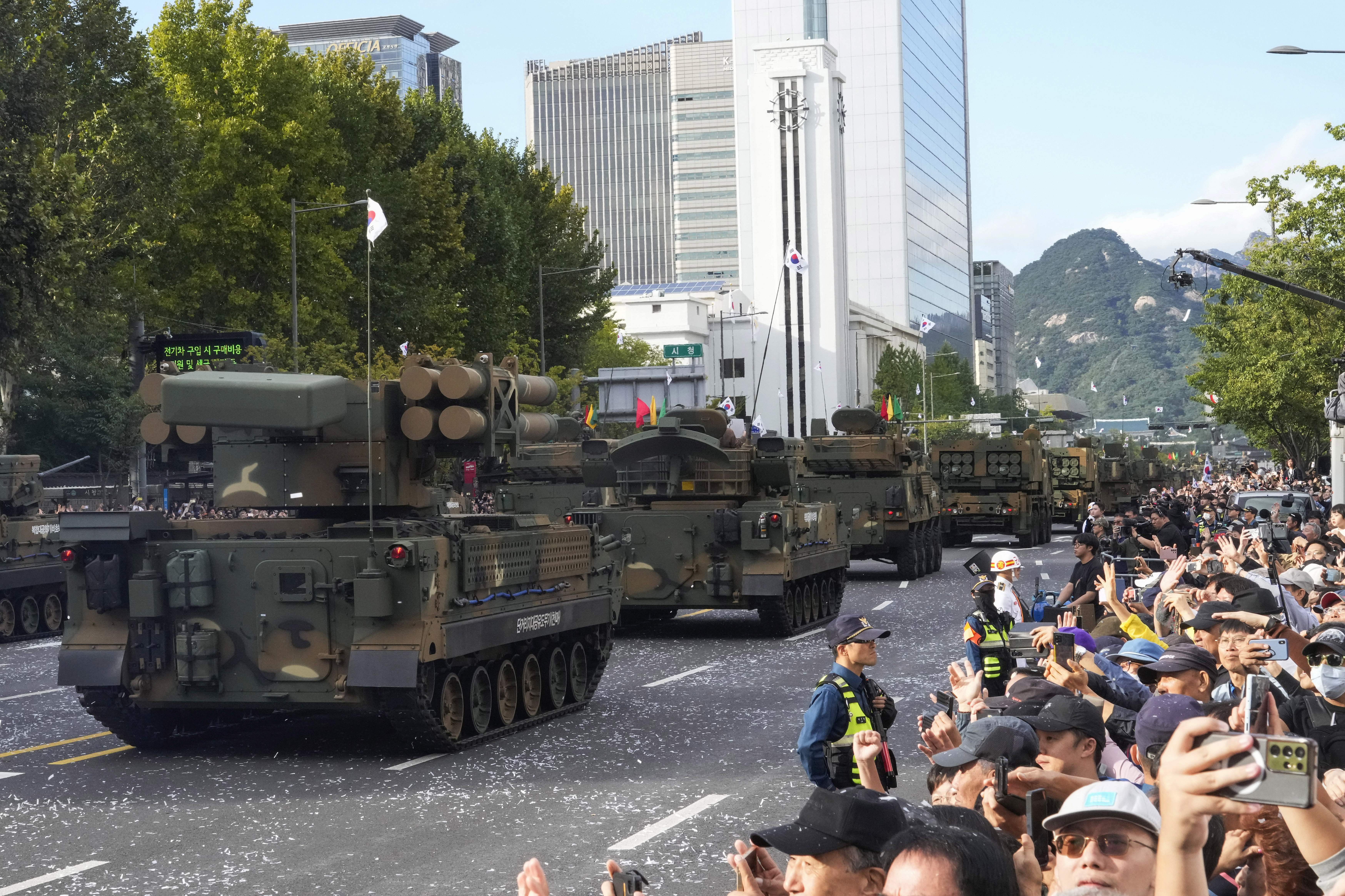 South Korean missiles driven through the streets of Seoul. There are tower blocks behind. A crowd of people along the road are waving and cheering.
