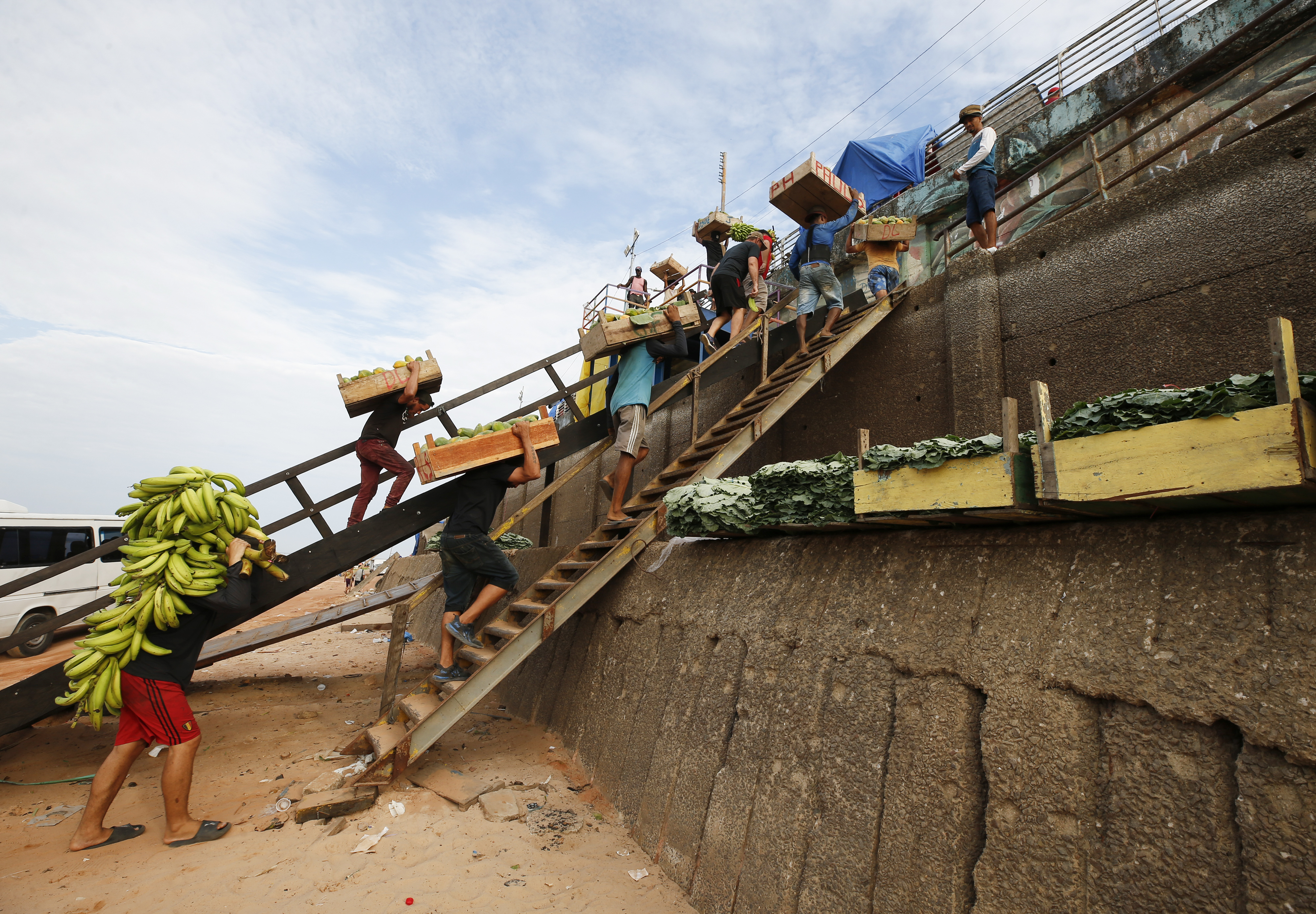 Workers move food like bananas and other goods down a wooden staircase onto a dry river bank, off a boat.