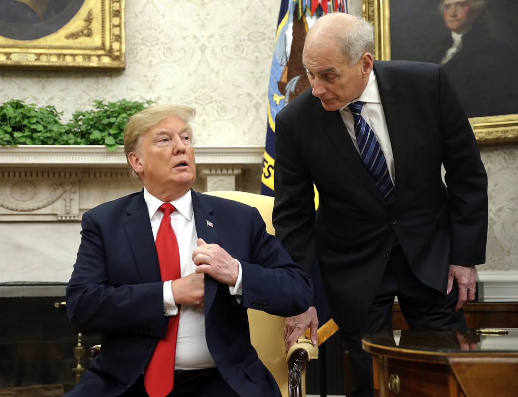John Kelly standing next to Trump, who is sitting in a chair and wearing a red necktie