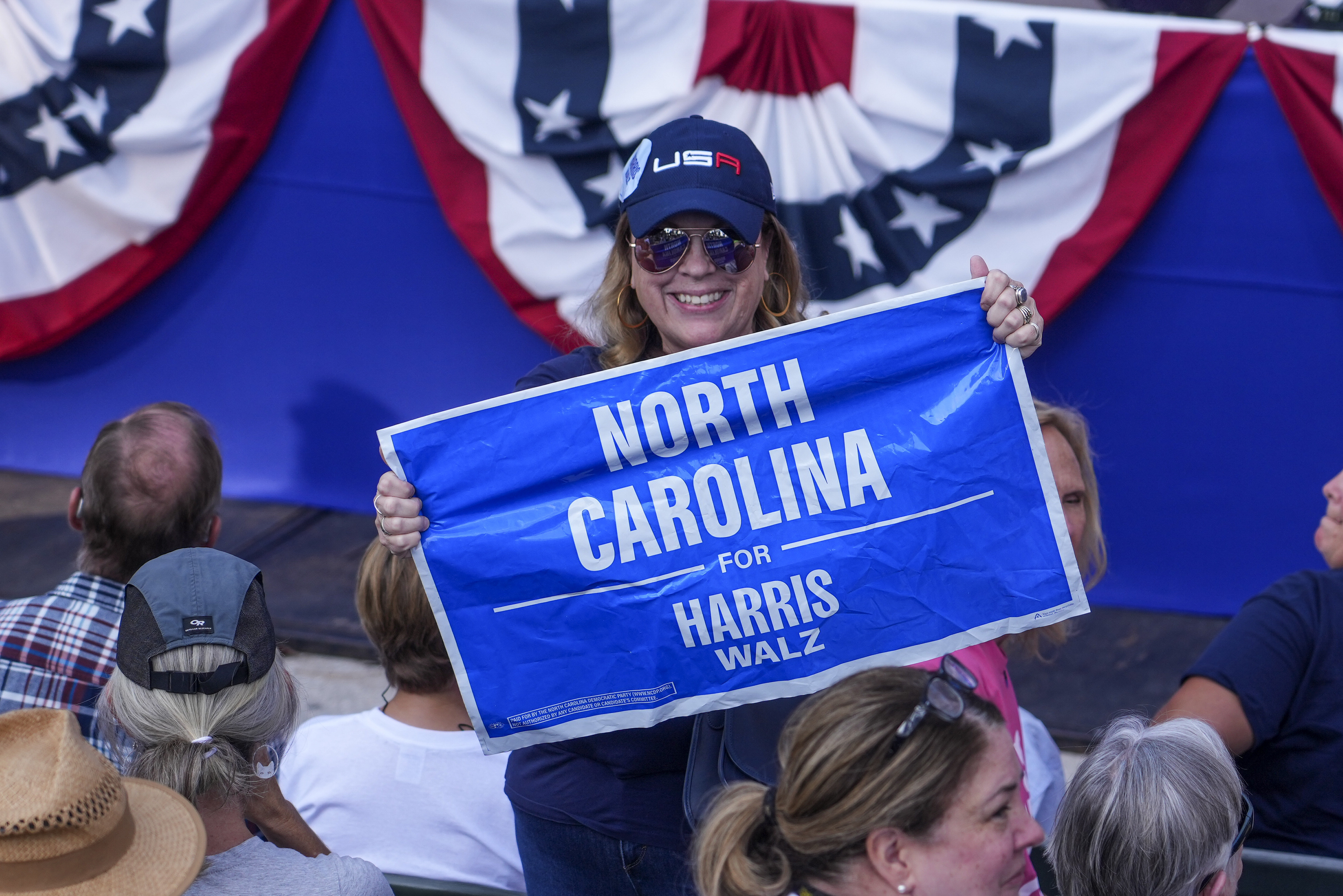 A woman in a USA baseball cap holds up a sign that reads, "North Carolina is for Harris Walz."