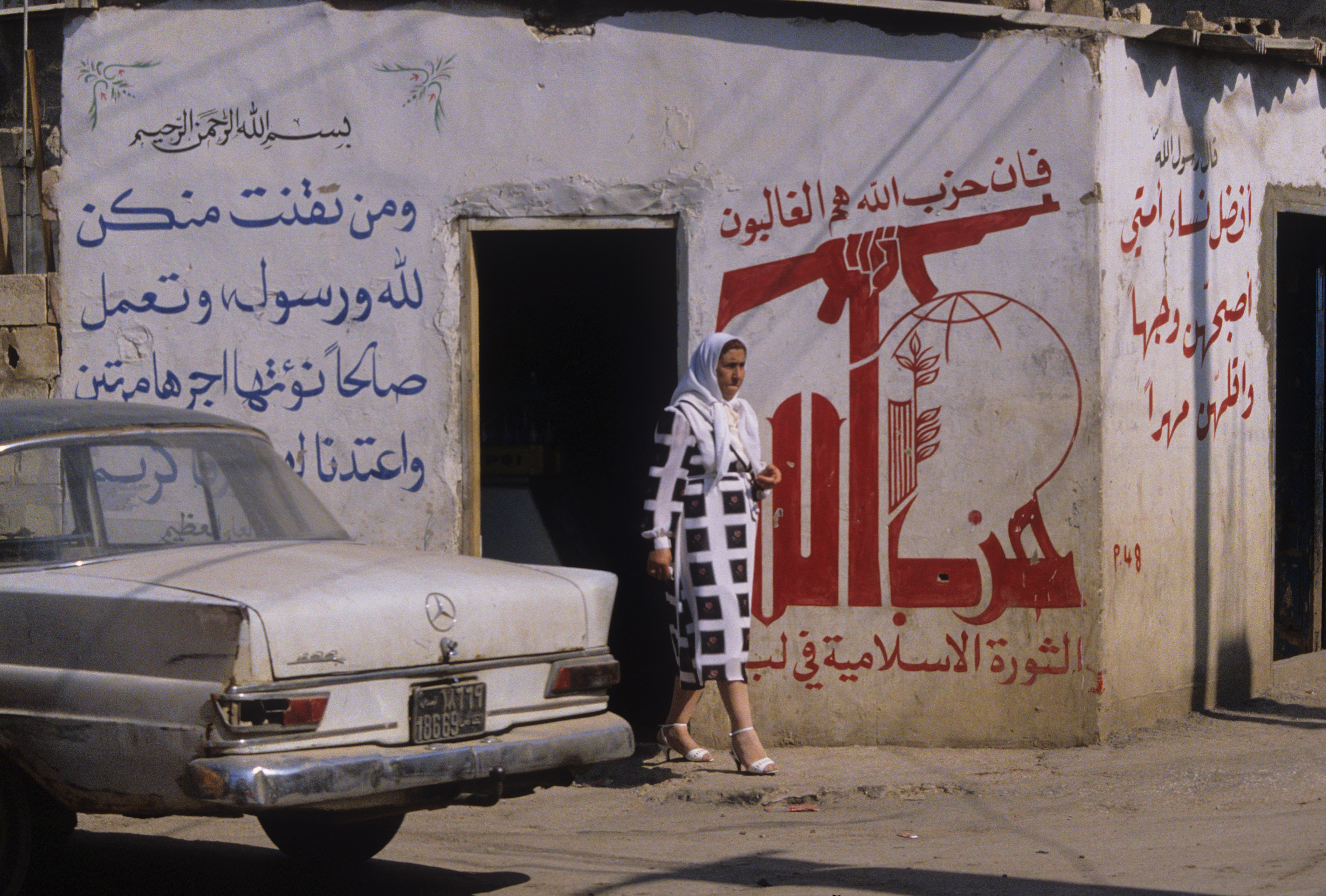 A woman in a veil walks past an old 1960s Mercedes Benz parked in front of a Hezbollah mural in southern suburb of Beirut during the civil war, 12th June 1986.