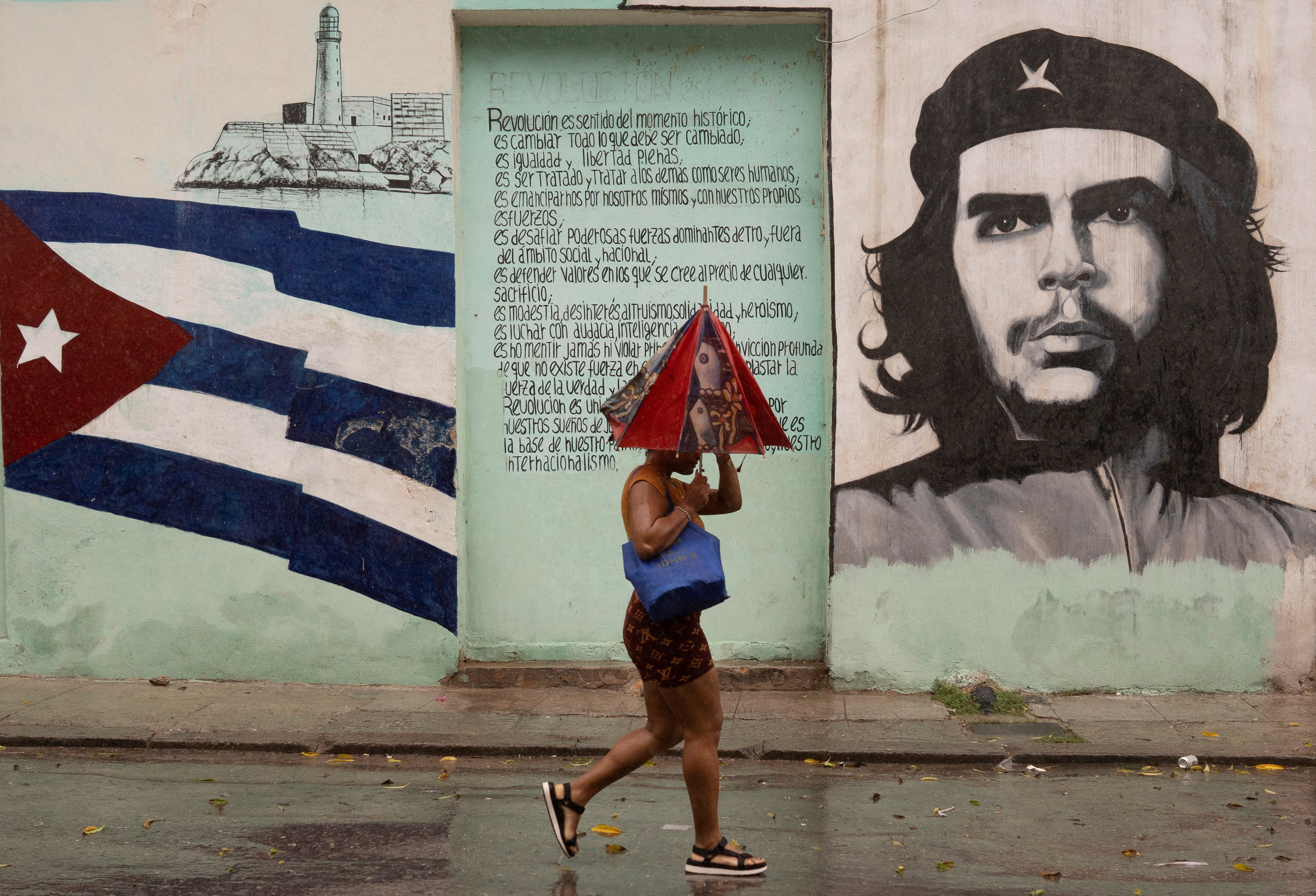 A person under a crumpled umbrella walks past murals of the Cuban flag and Che Guevara