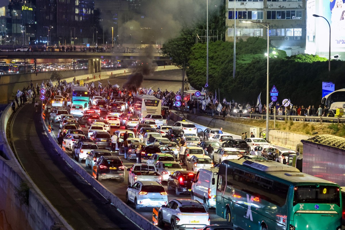 Cars are jammed together on a highway, unable to move further as a line of protesters stands in front with Israeli flags. It's nighttime.