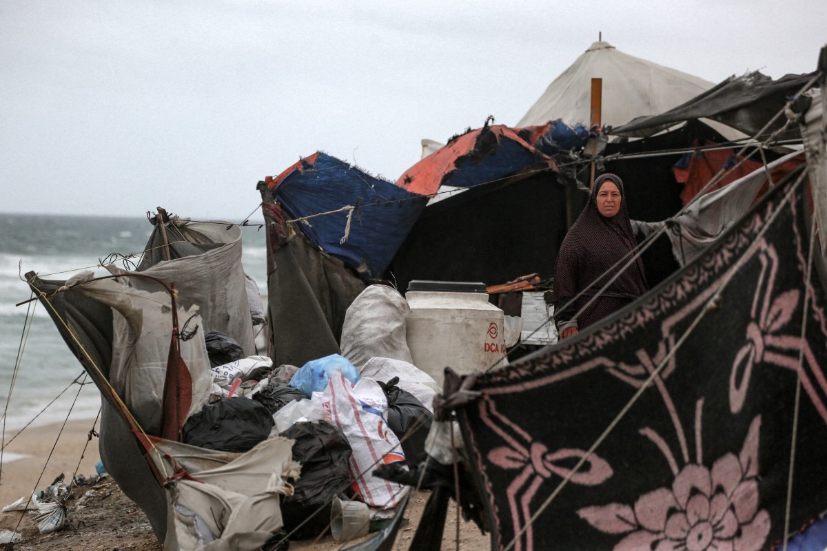 A displaced Palestinian woman stands inside a wind and rain-damaged tent,