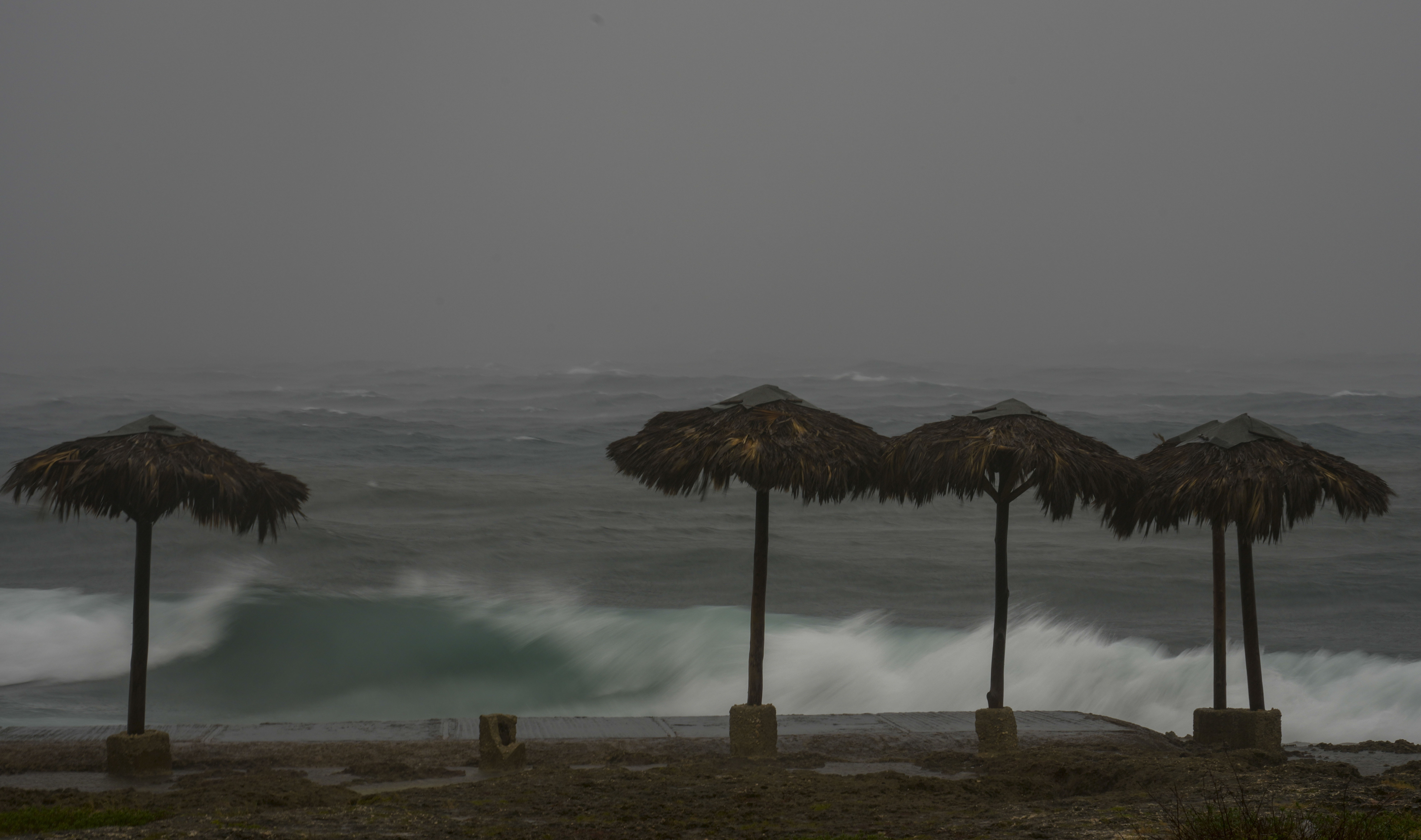 Round shades made of palm fronds stand on a beach in front of stormy seas.