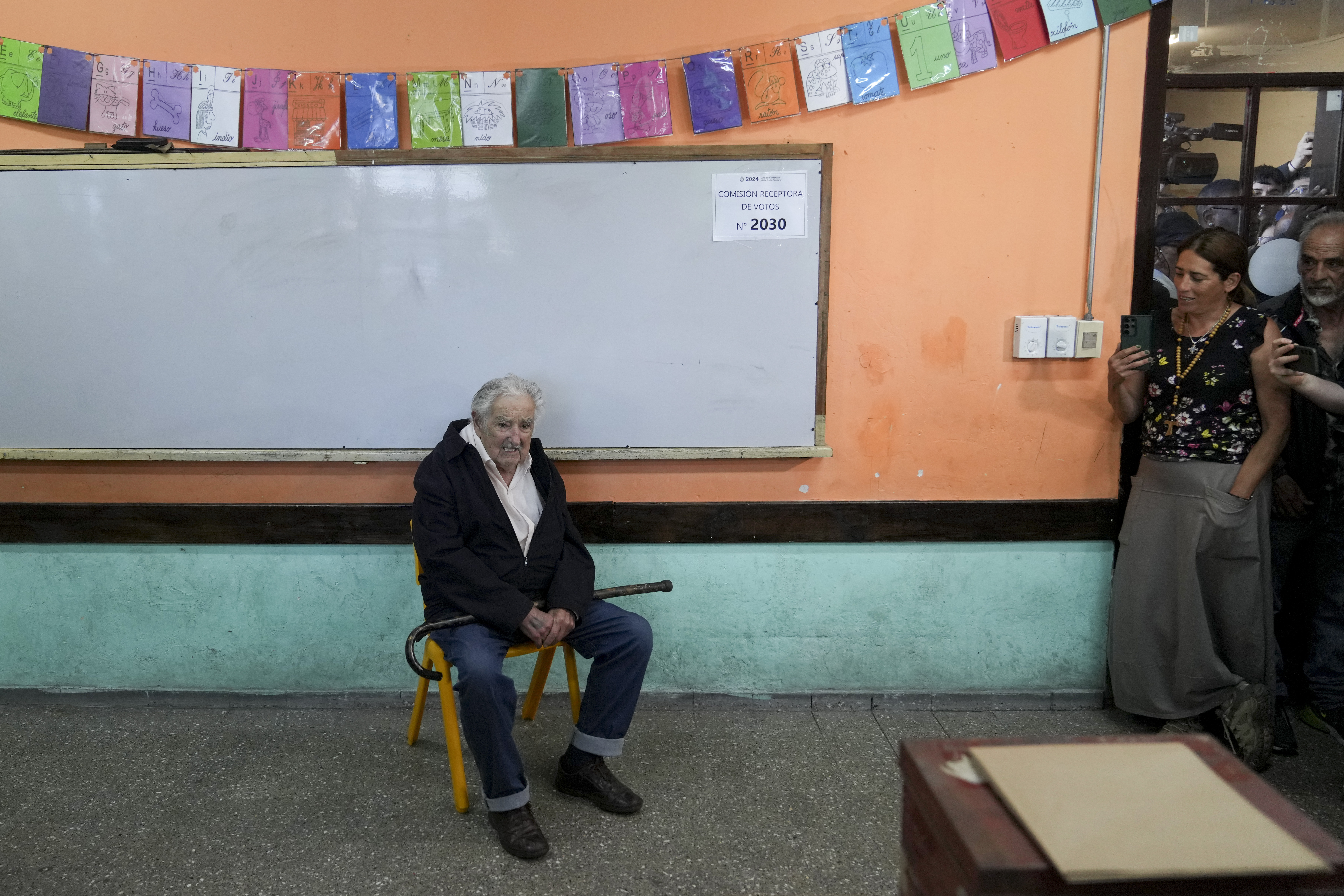 Mujica sits on a chair with his walking stick on his lap. The room is a classroom with a large white board behind him.