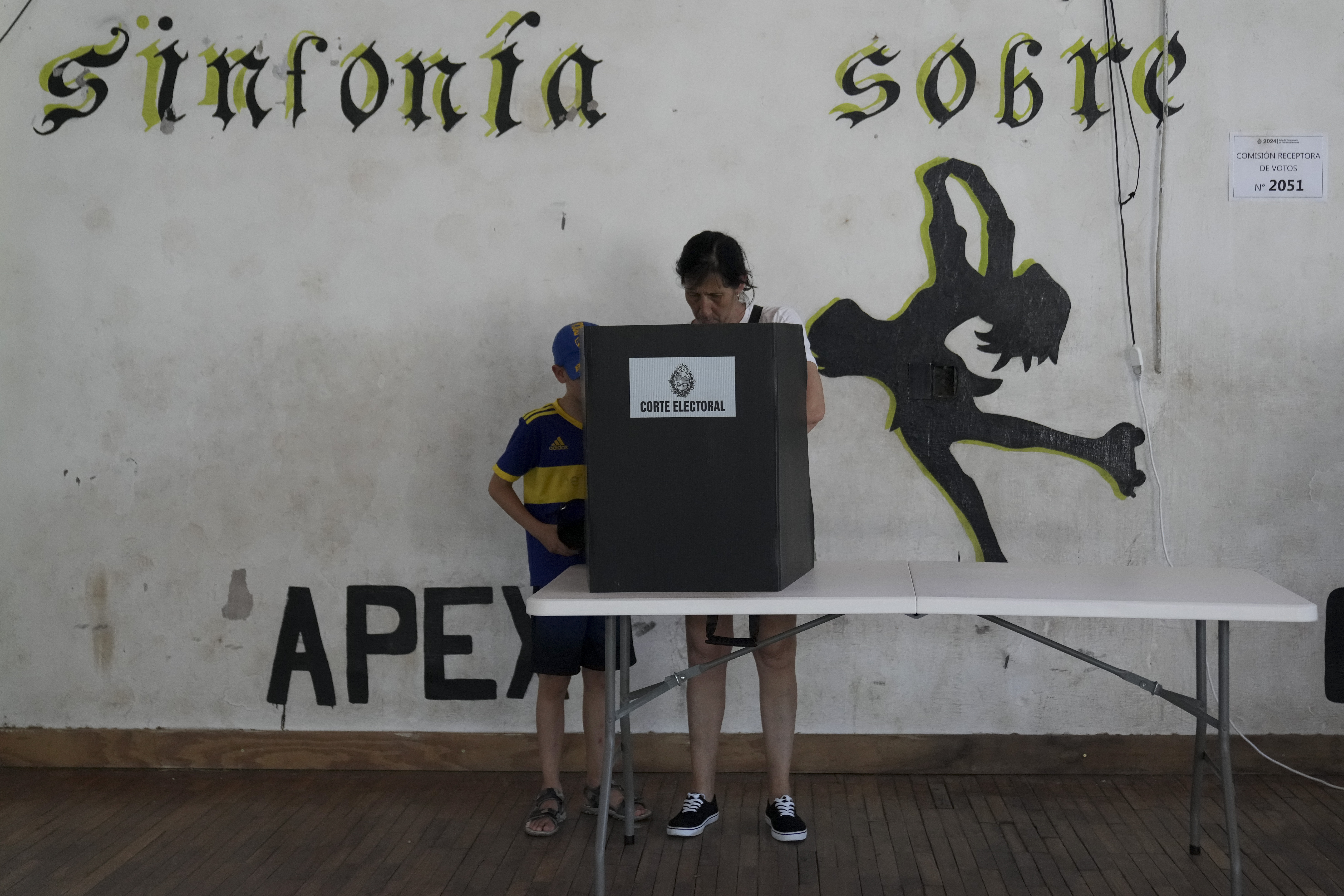 A voter, standing in front of a wall marked with murals and slogans, casts a ballot behind a privacy screen with their child.