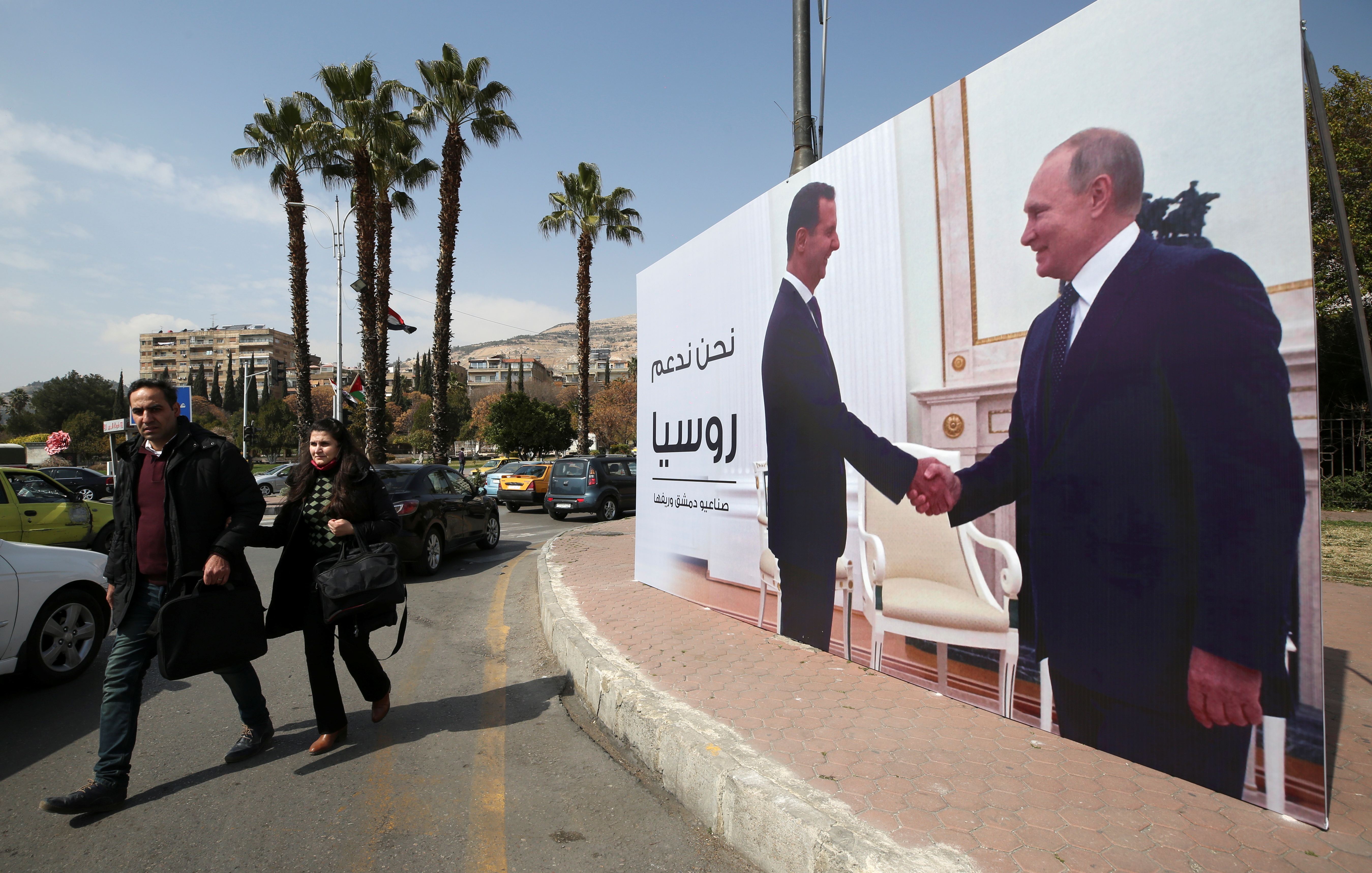 People walk past a poster depicting Syrian President Bashar al-Assad shaking hands with Russian President Vladimir Putin in Damascus, Syria