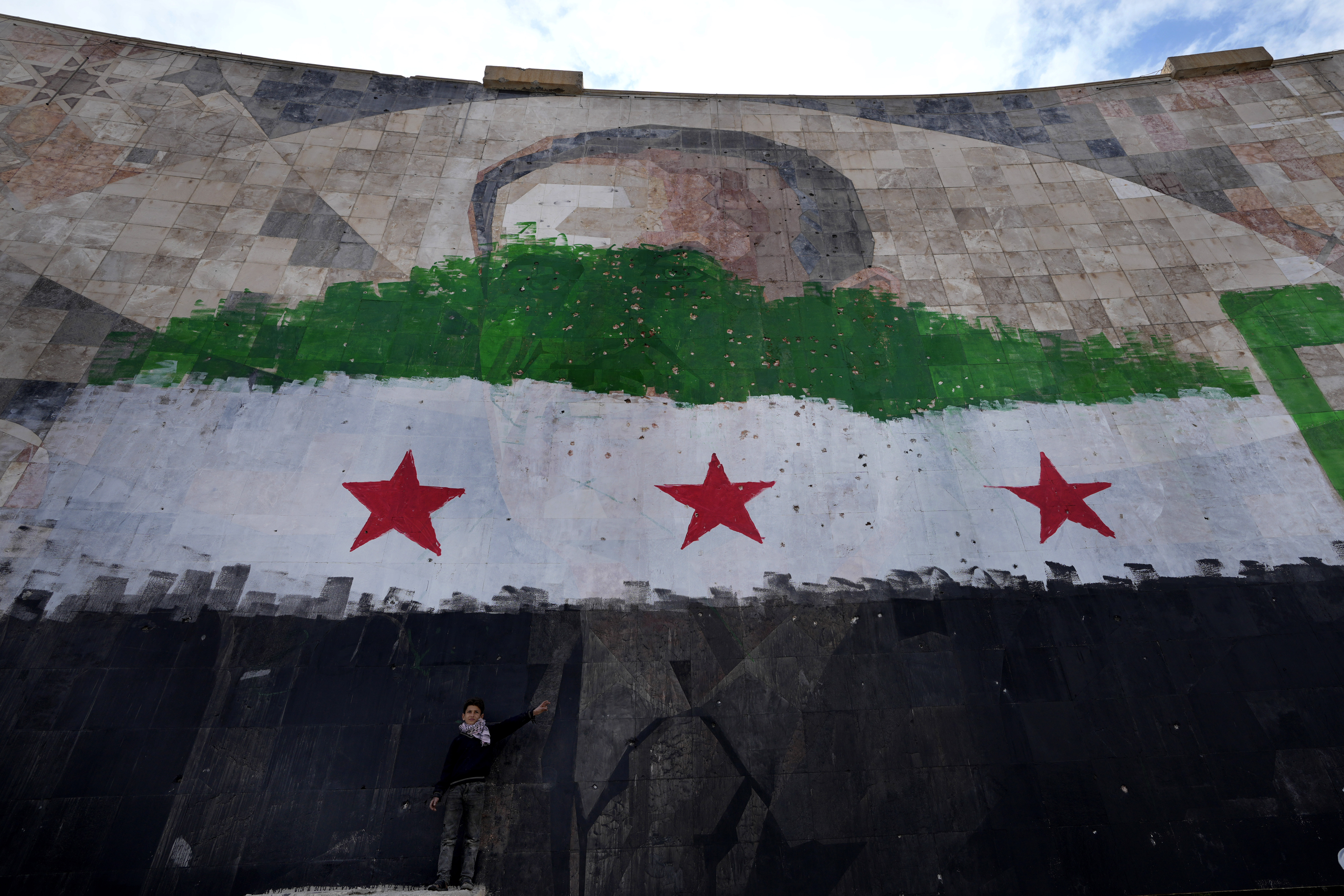 A Syrian boy stands under a giant portrait of the late Syrian President Hafez Assad painted over with the colors of the "revolutionary" flag, in Damascus, Syria, Thursday, Dec. 26