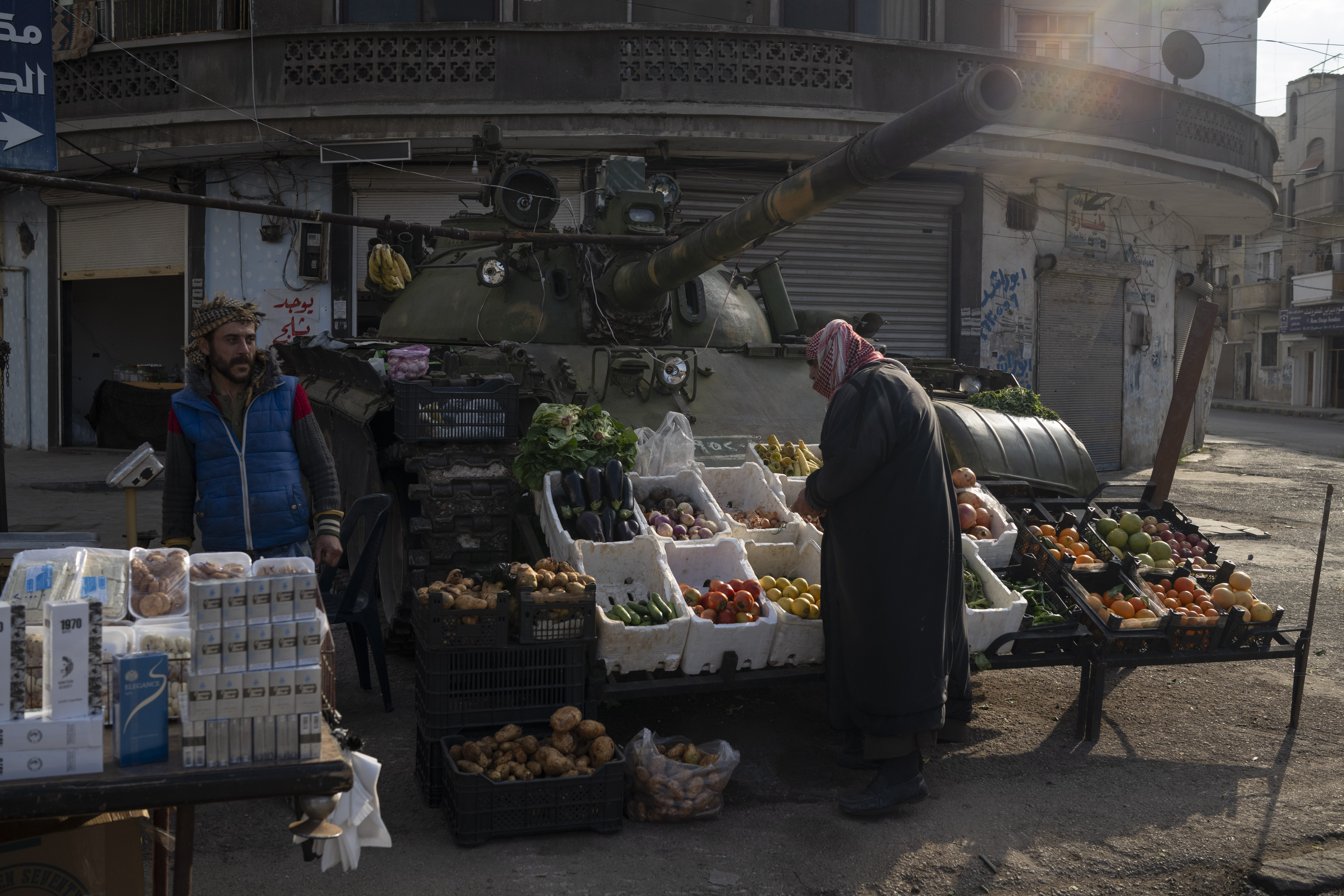 A man looks at fruits and vegetables displayed for sale in front of an ousted Syrian government forces tank that was left on a street in an Alawite neighbourhood, in Homs, Syria, Thursday, Dec. 26