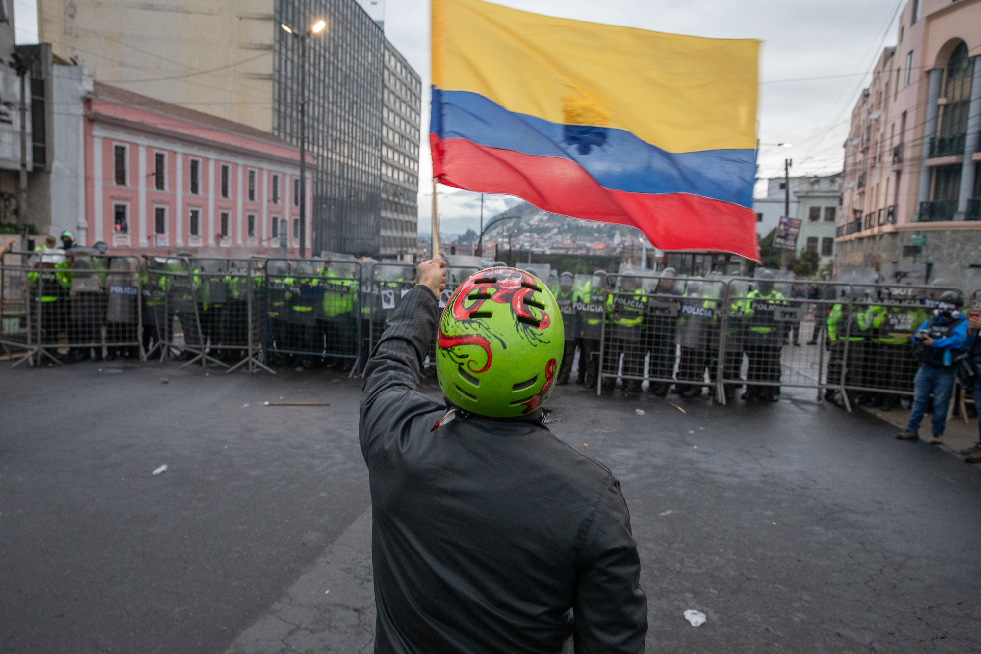 A protester in a green helmet waves an Ecuadorian flag in front of a line of police