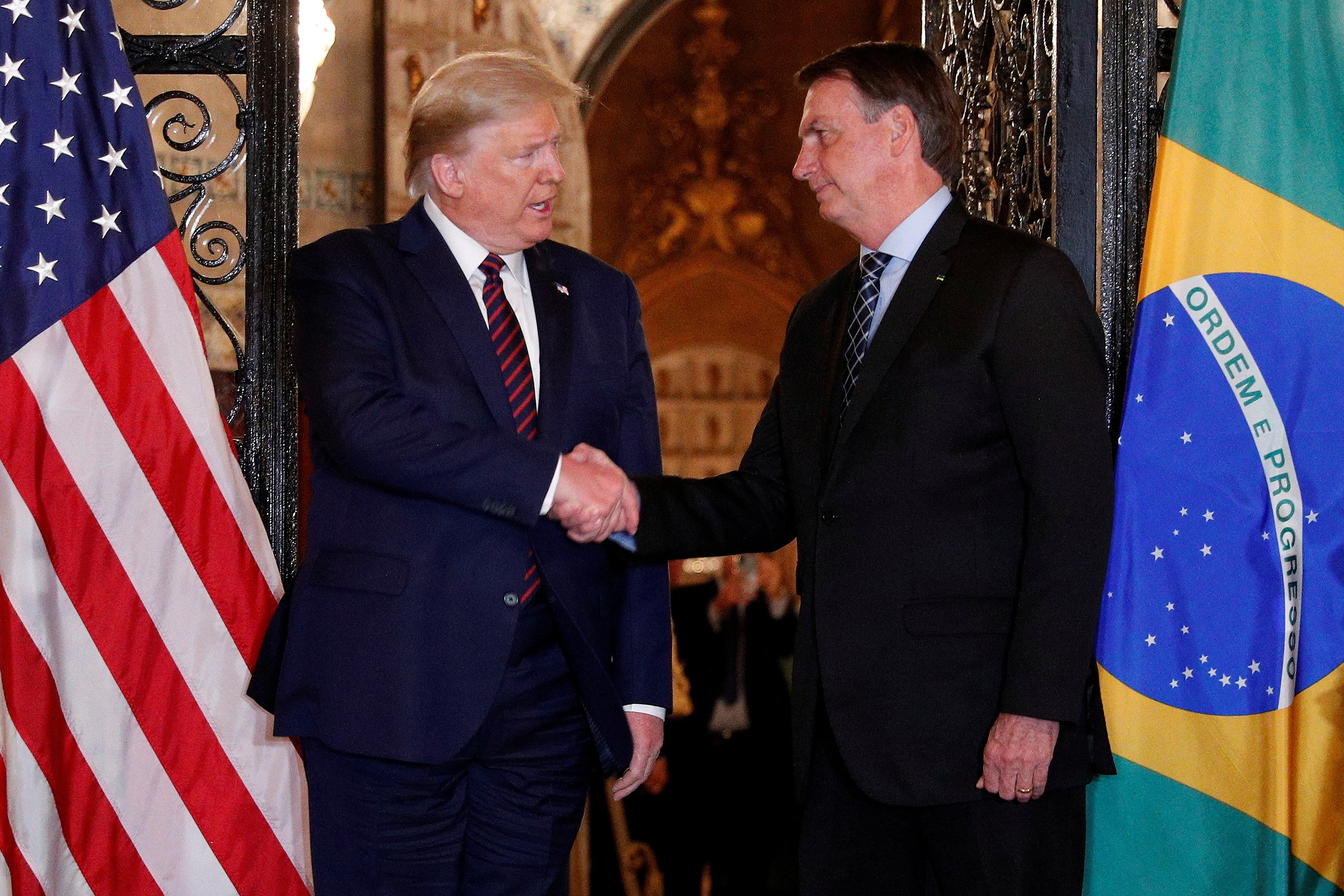 Donald Trump shakes Jair Bolsonaro's hand as they stand between their two countries' flags