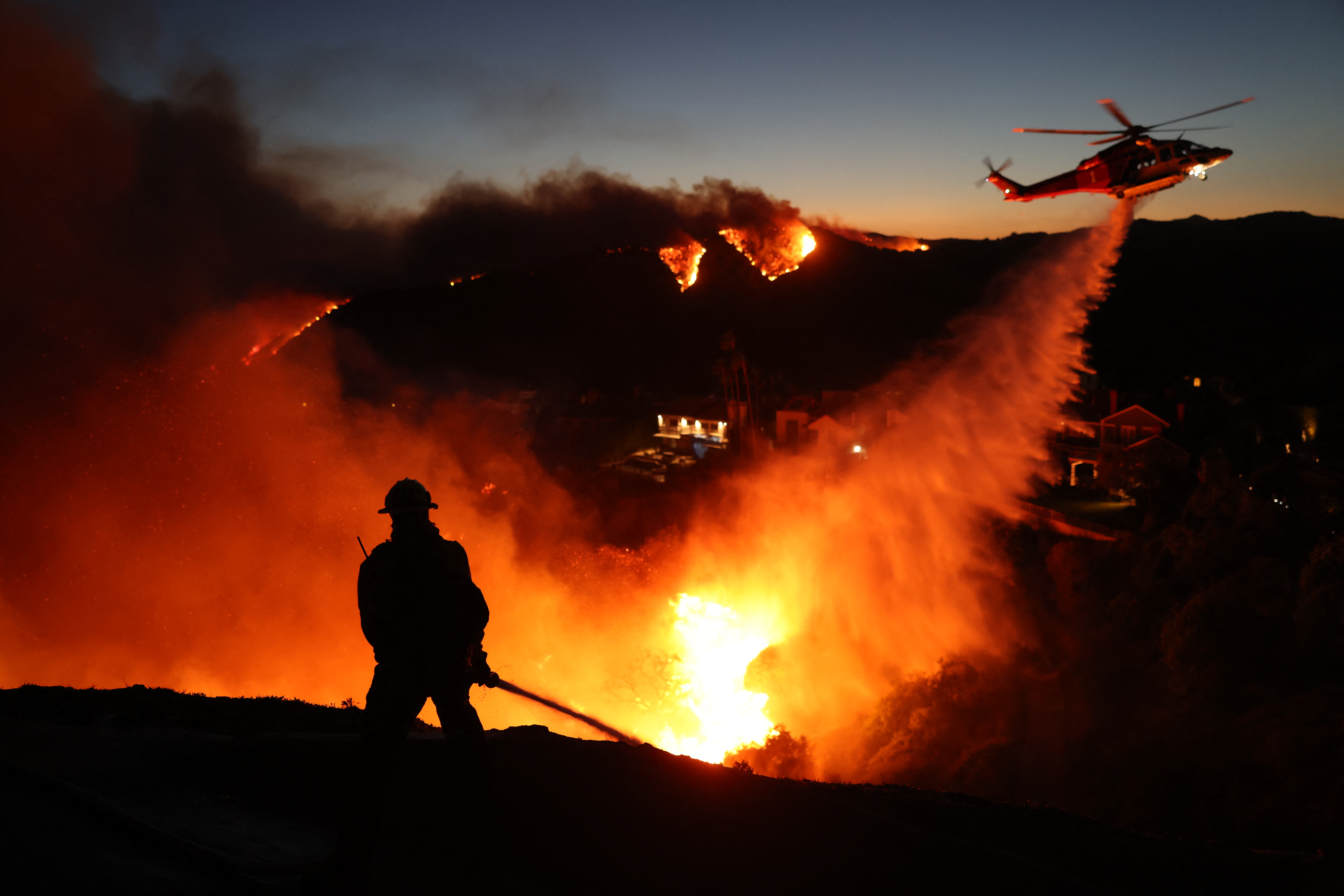 Fire personnel respond to homes destroyed while a helicopter drops water as the Palisades Fire grows in Pacific Palisades, California on January 7