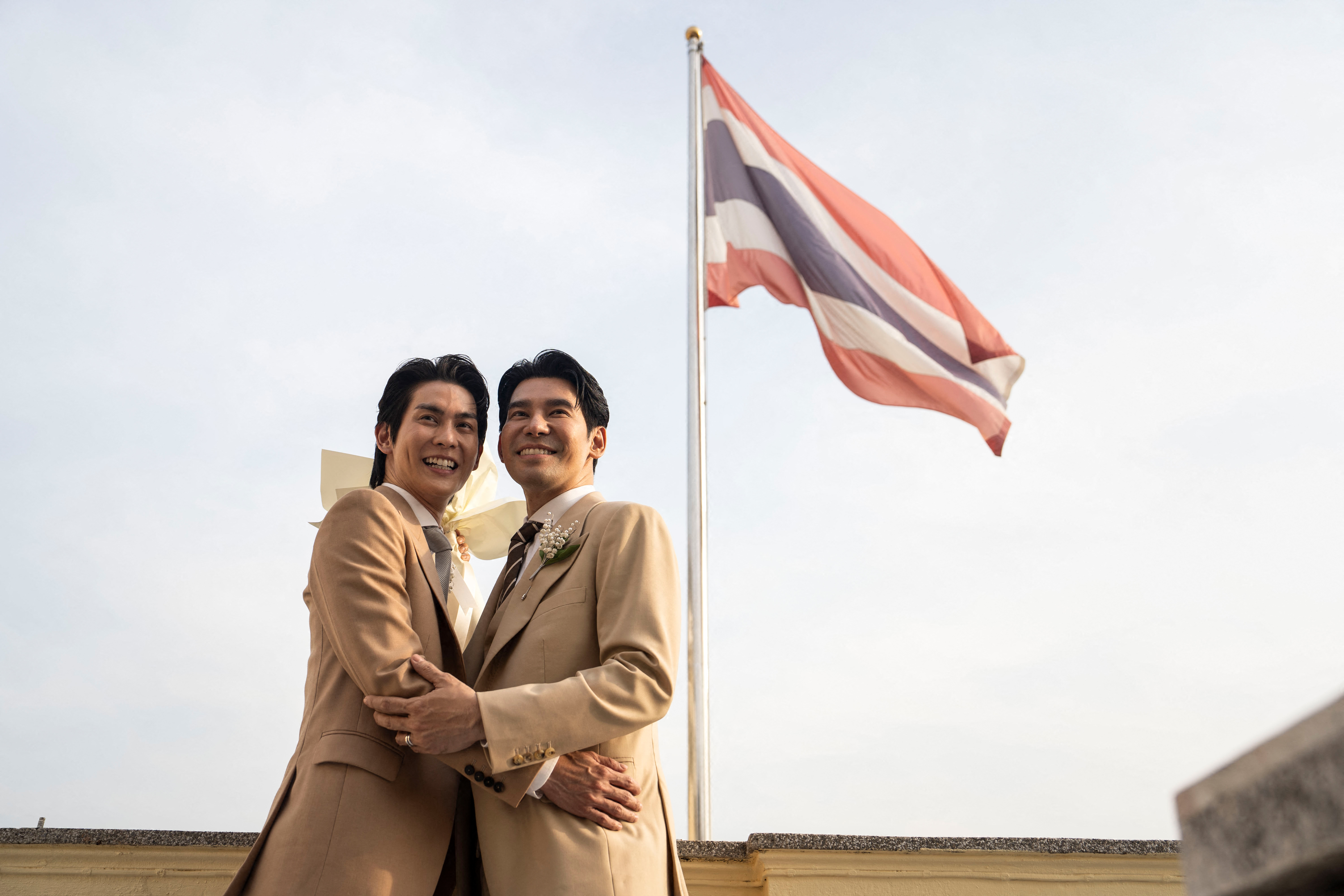 Thai actors Apiwat "Porsch" Apiwatsayree (R) and Sappanyoo "Arm" Panatkool pose for pictures in front of a Thai national flag after registering their same-sex marriage at the Phra Nakhon district office in Bangkok on January 23, 2025. A high-profile gay couple married in Thailand on January 23 as the kingdom's same-sex marriage law went into effect, an AFP journalist saw, among the first of hundreds expected to do so. (Photo by Chanakarn LAOSARAKHAM / AFP)