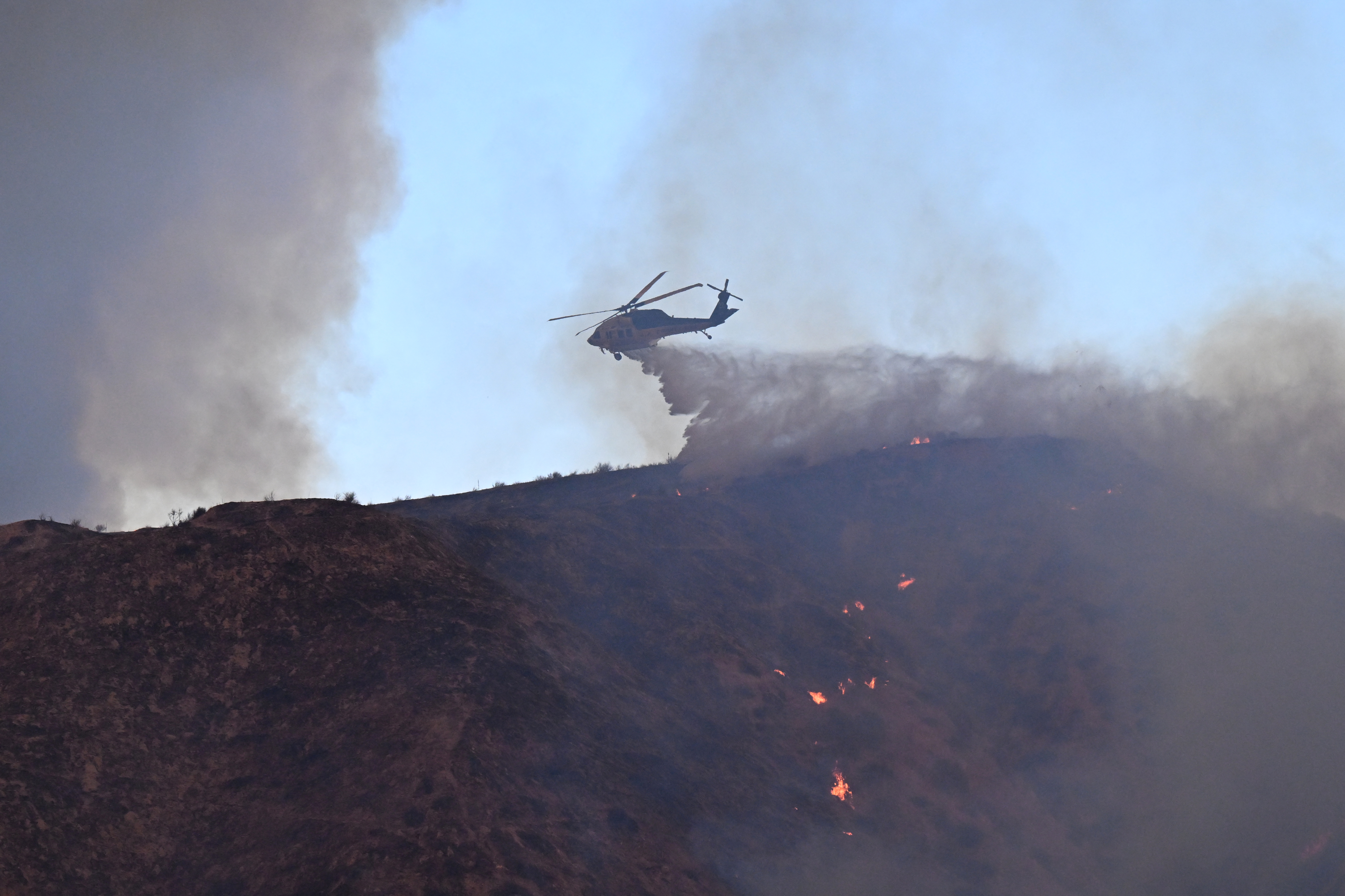 A firefighting helicopter drops water on the Hughes Fire in Castaic, a northwestern neighborhood of Los Angeles, California, January 22, 2025. A new wildfire erupted north of Los Angeles on January 22, exploding in size and sparking thousands of evacuation orders in a region already staggering from the effects of huge blazes. Ferocious flames were devouring hillsides near Castaic Lake, spreading rapidly to cover 5,000 acres (2,000 hectares) in just over two hours. The fire was being fanned by strong, dry Santa Ana winds that were racing through the area, pushing a vast pall of smoke and embers ahead of the flames. Evacuations were ordered for 19,000 people all around the lake, which sits around 35 miles north of Los Angeles, and close to the city of Santa Clarita. (Photo by Robyn Beck / AFP)