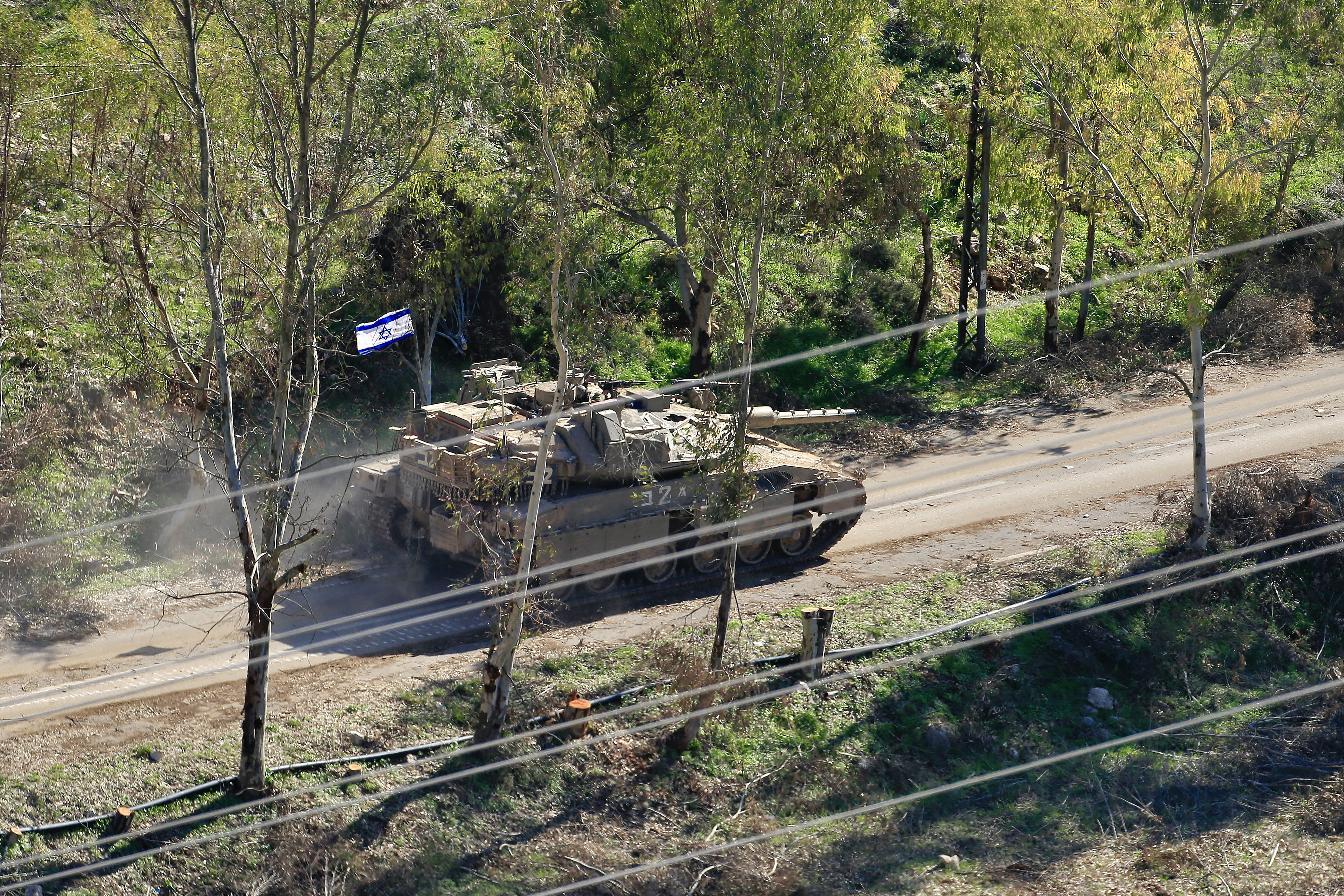 A tank with an Israeli flag moves on an unpaved road between dense green trees.