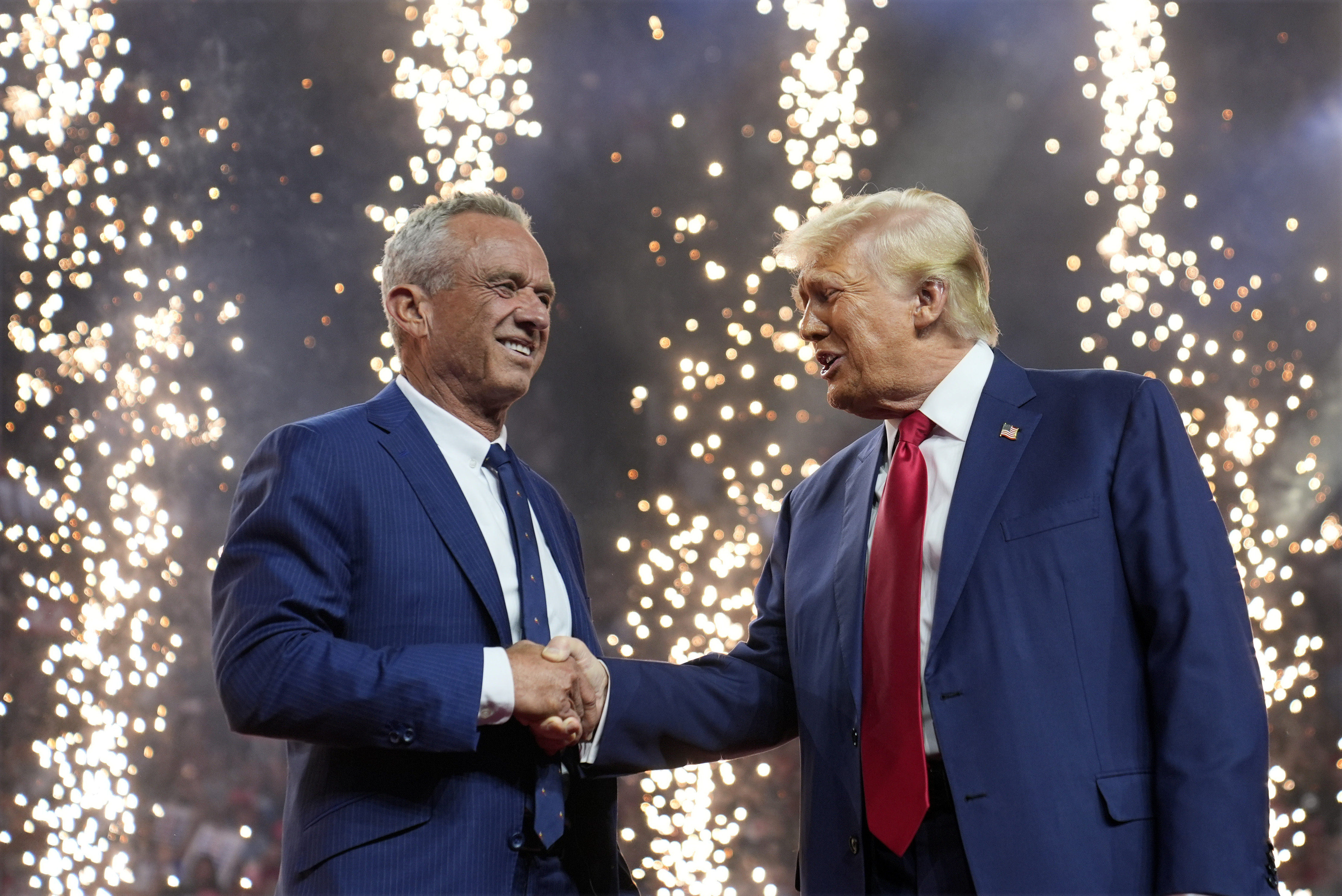 Donald Trump shakes hands with Robert F Kennedy Jr in front of a festive waterfall of sparks.