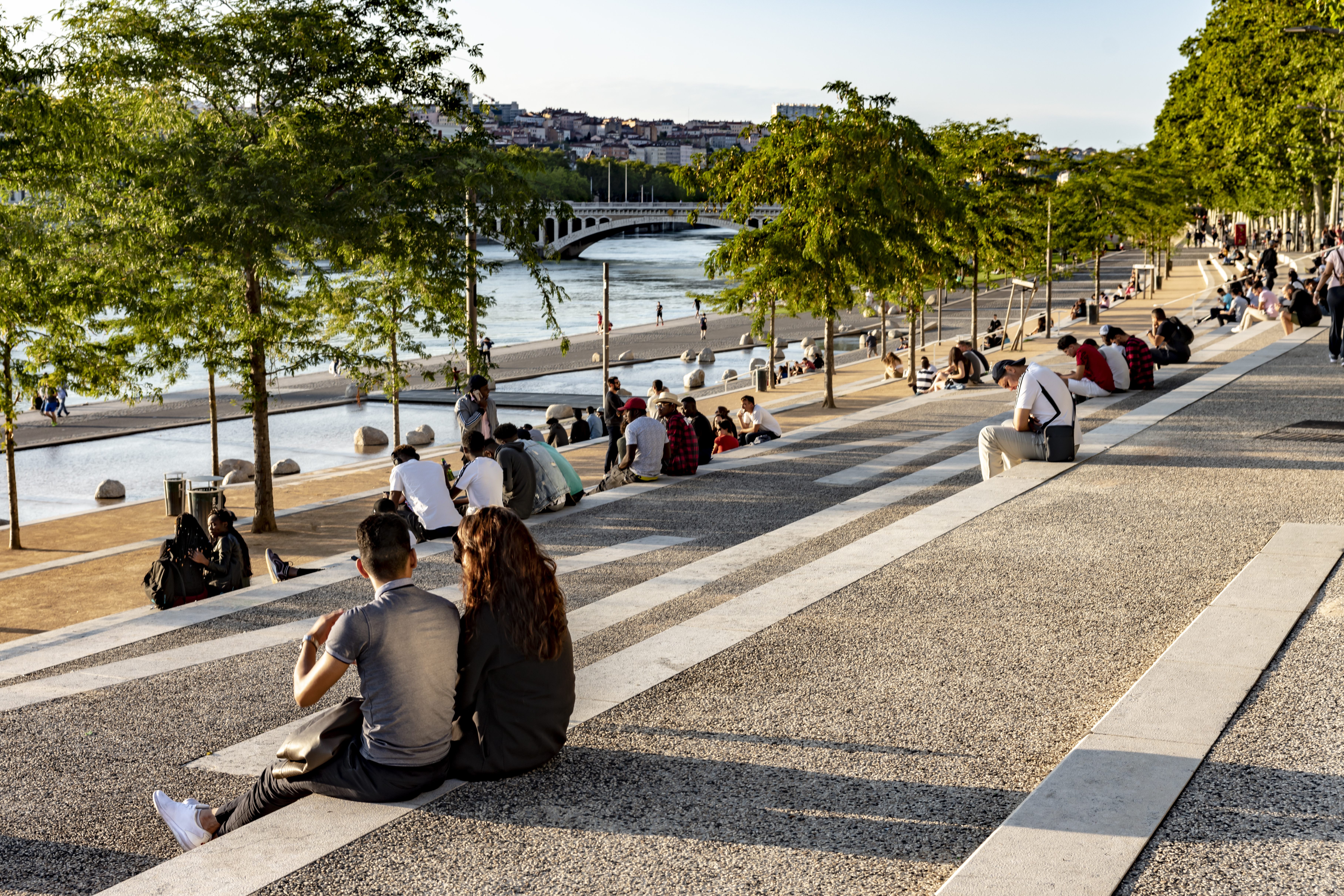 The banks of the Rhone at Lyon. There are groups of young people sitting on the steps beside the promenade along the banks of the river.