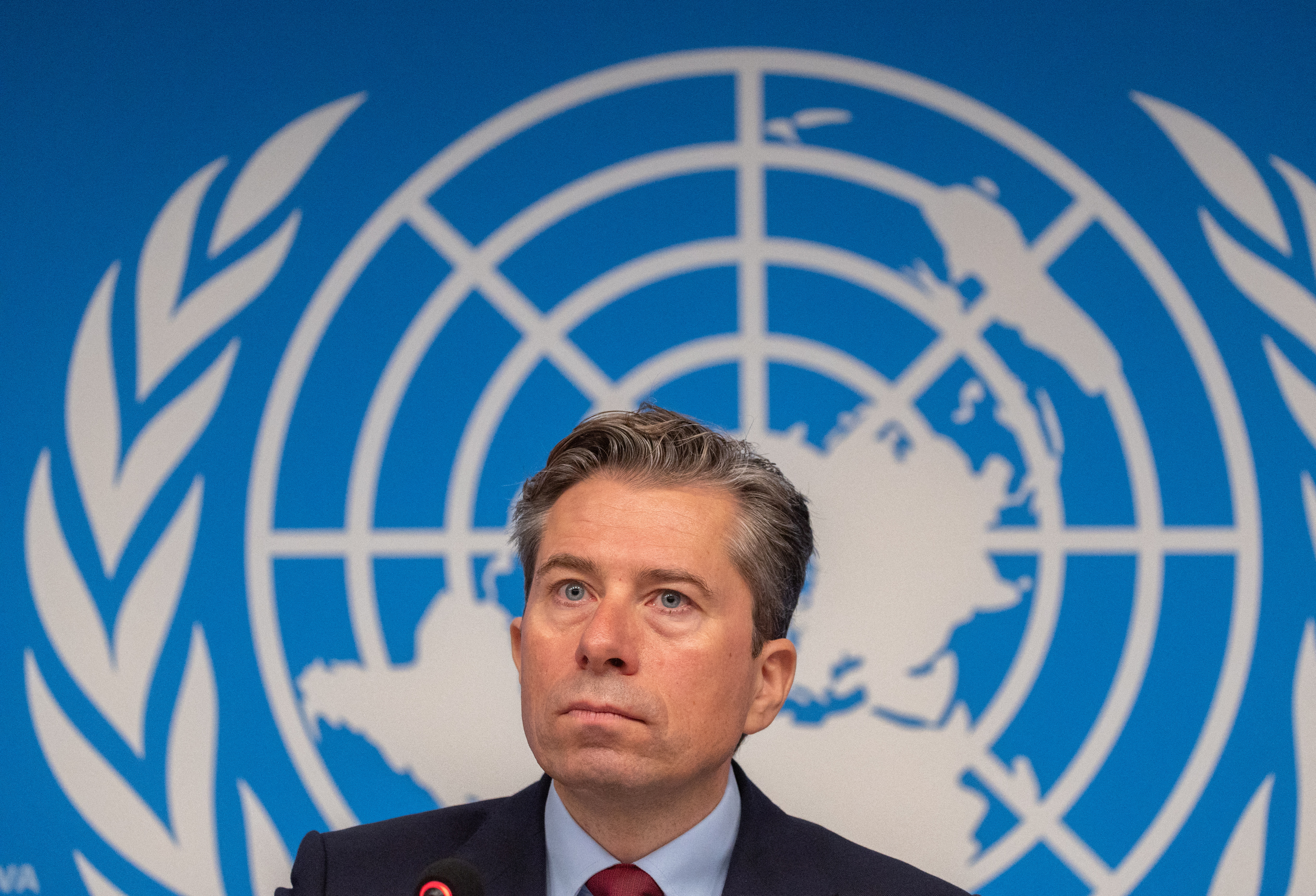 a man in a suit sits in front of a UN emblem on a blue background