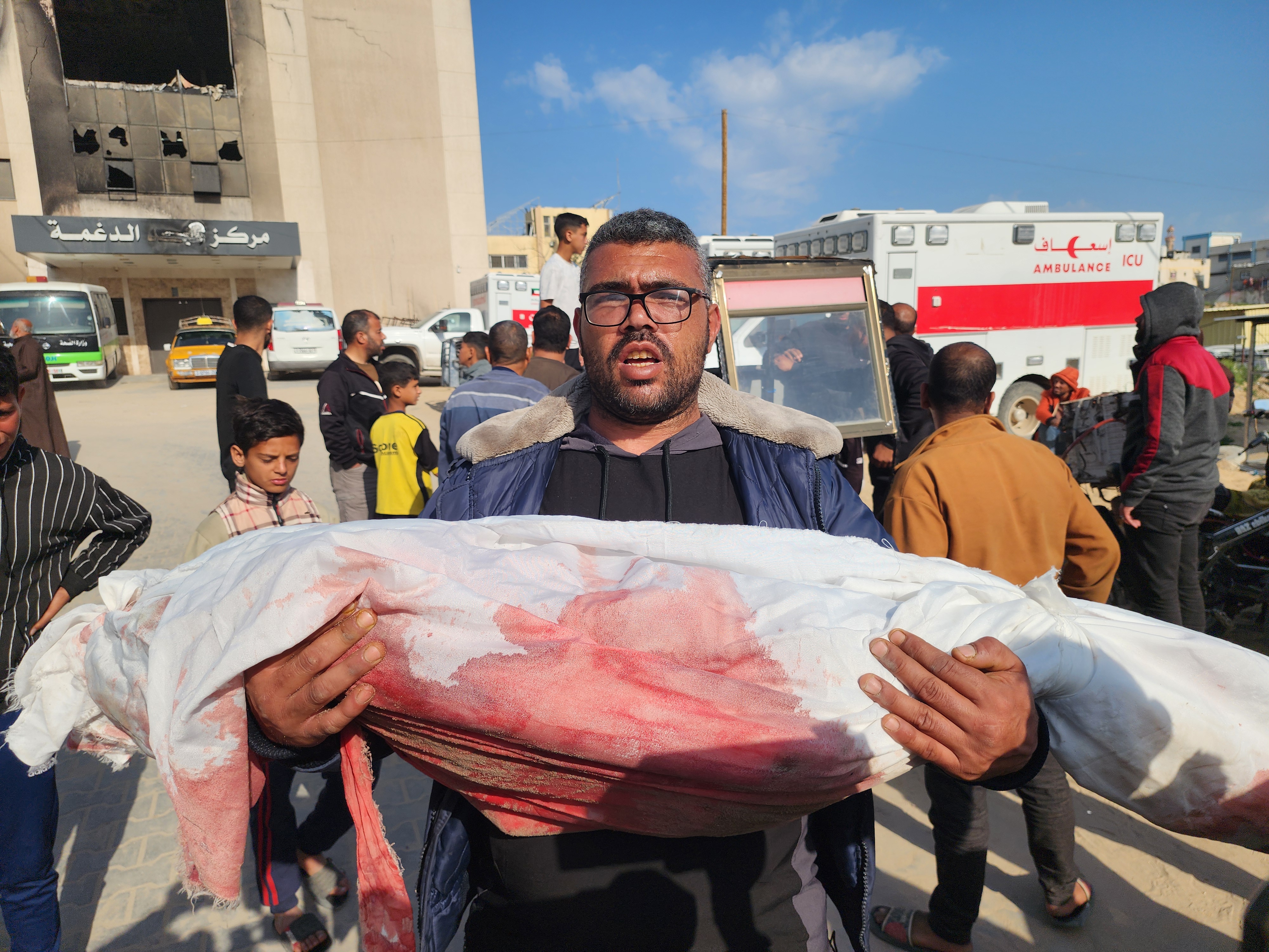 Man carrying the bloodstained, shrouded body of a child through the streets of Gaza