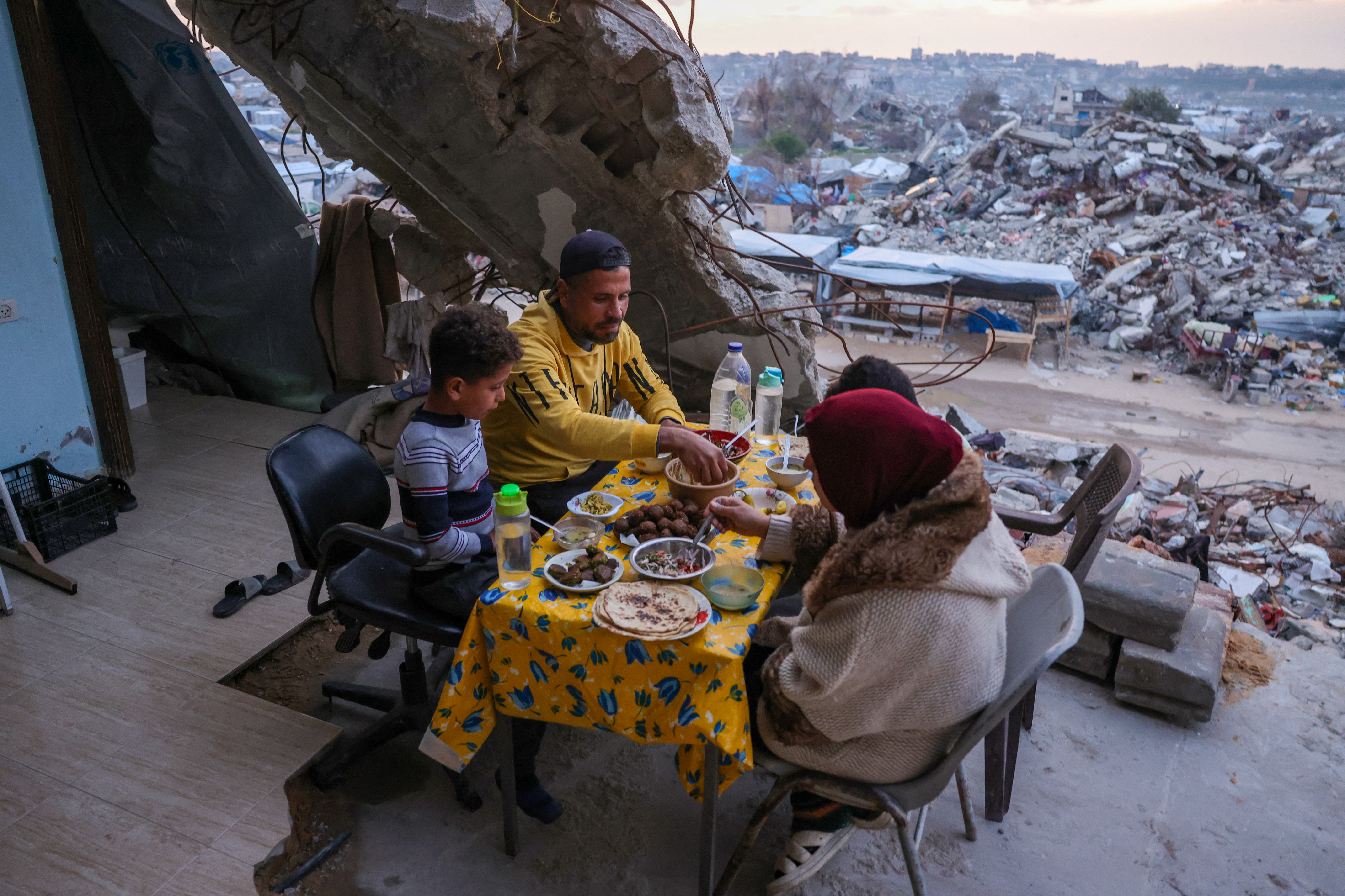 a family sit around a table with a table cloth next to destroyed buildings