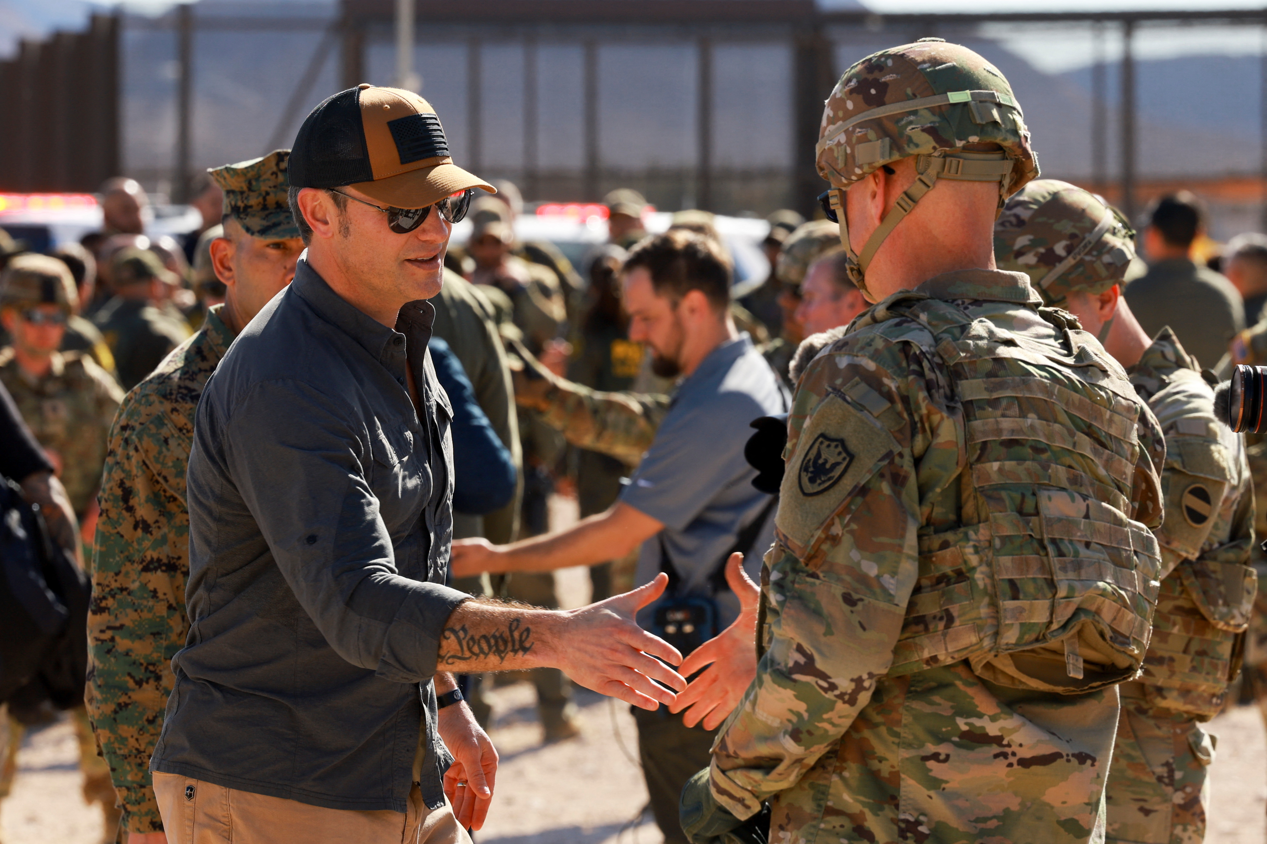 Pete Hegseth, in a ballcap and sunglasses, shakes hands with a soldier in fatigues and a helmet at the US southern border.