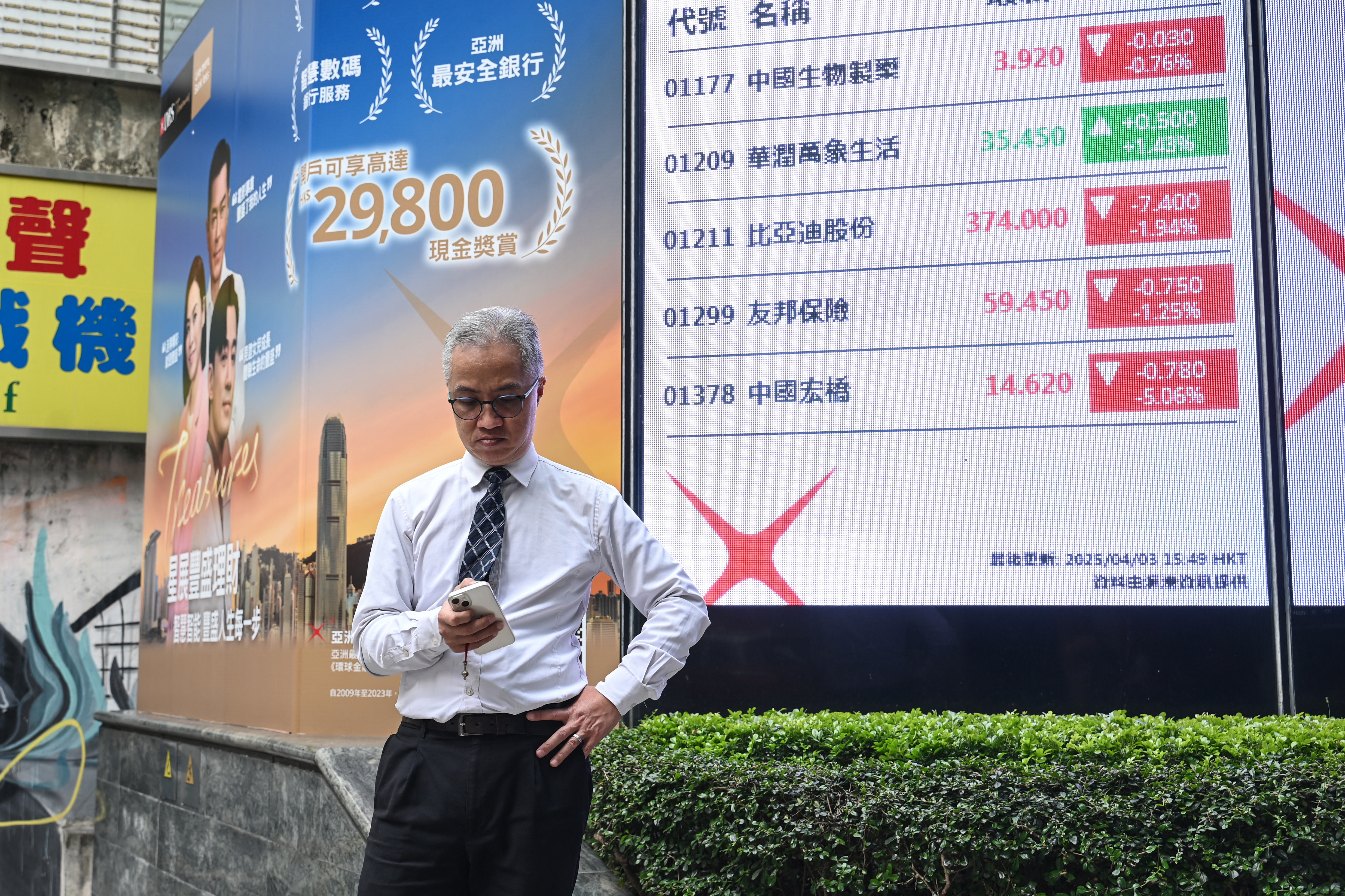 A man checks his phone next to an electronic board showing stocks on the Heng Seng Index in Hong Kong