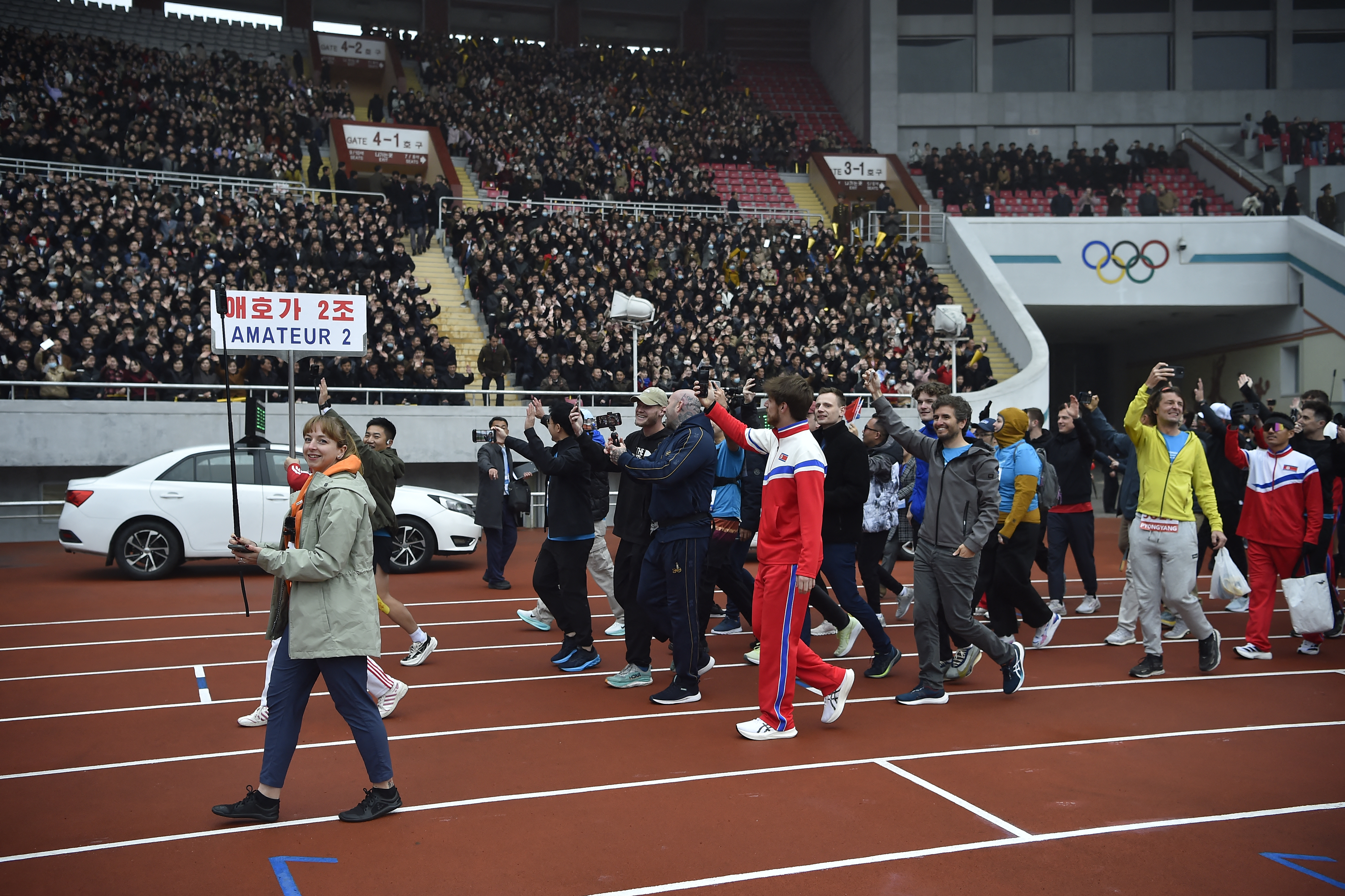 People walk along the racetrack, some wearing tracksuits. They take photos and smile as the crowd on the stands watches on.