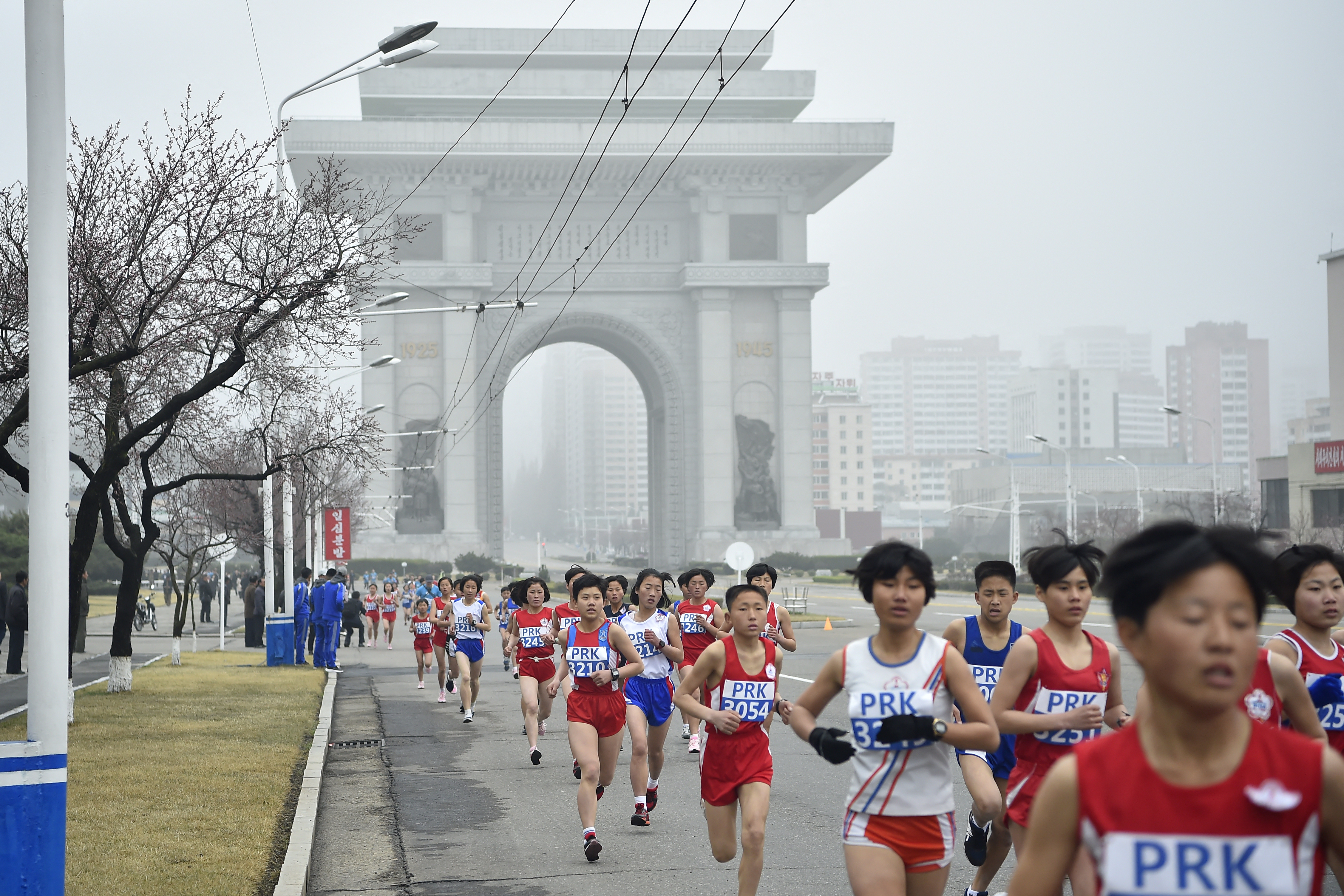 Athletes in red and blue run along the road with Pyongyang's white stone Arch of Triumph behind them.