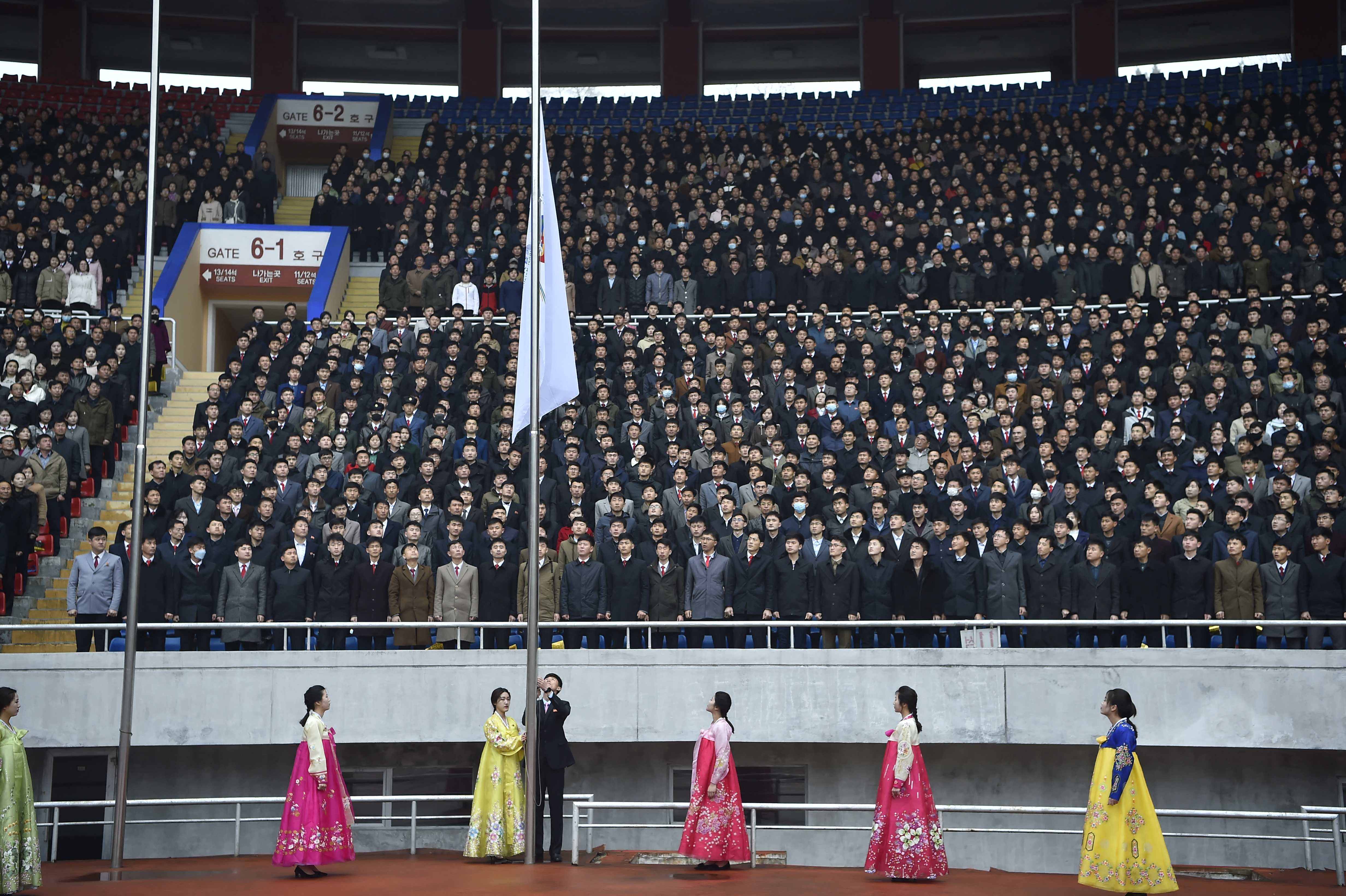 Women in beautiful, ceremonial dresses stand on the track as a man raises a flag and an audience watch on from the stands. Audience members are dressed in suits.