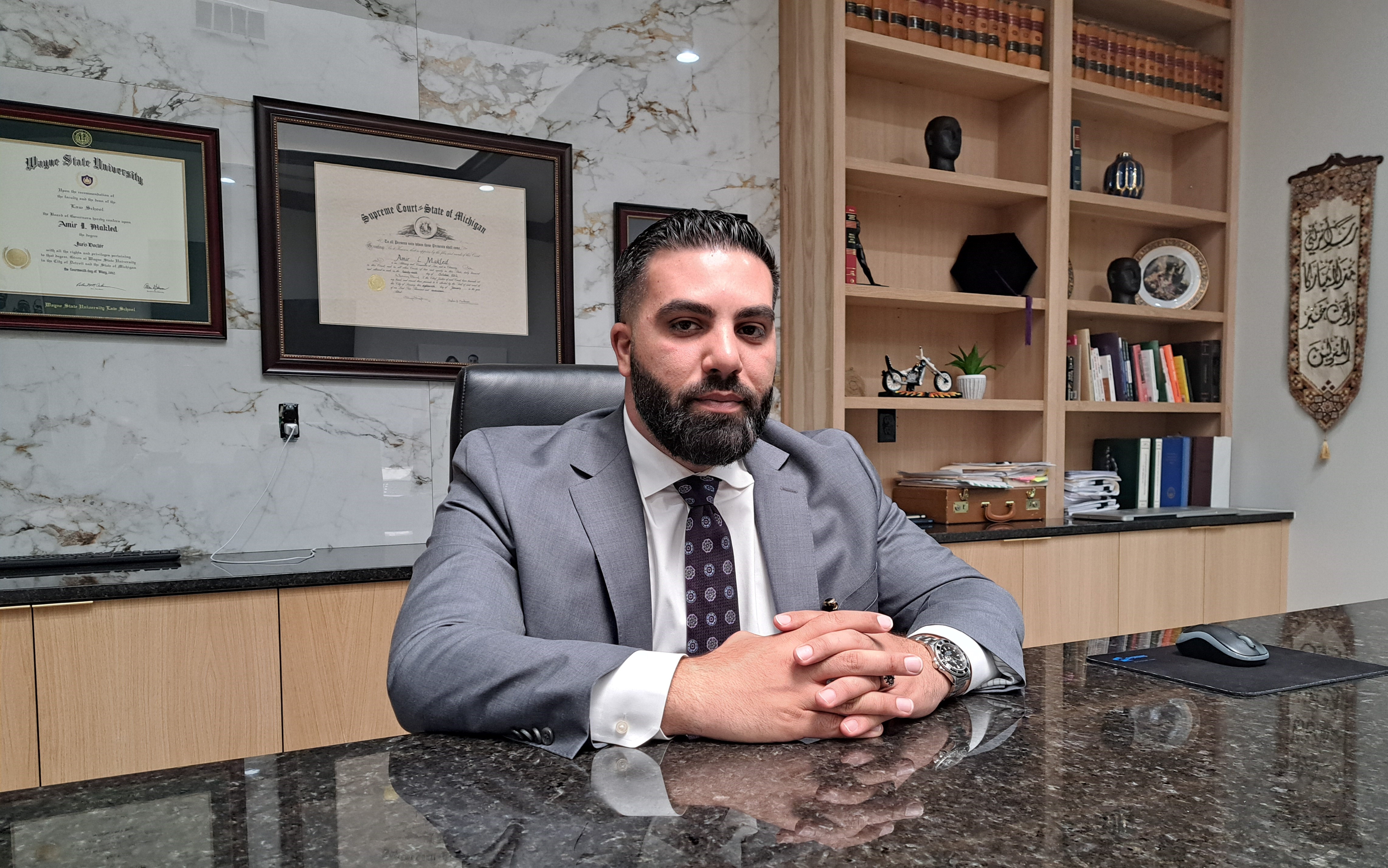 Amir Makled sits at a table with his hands folded, a law degree visible on the wall behind him.