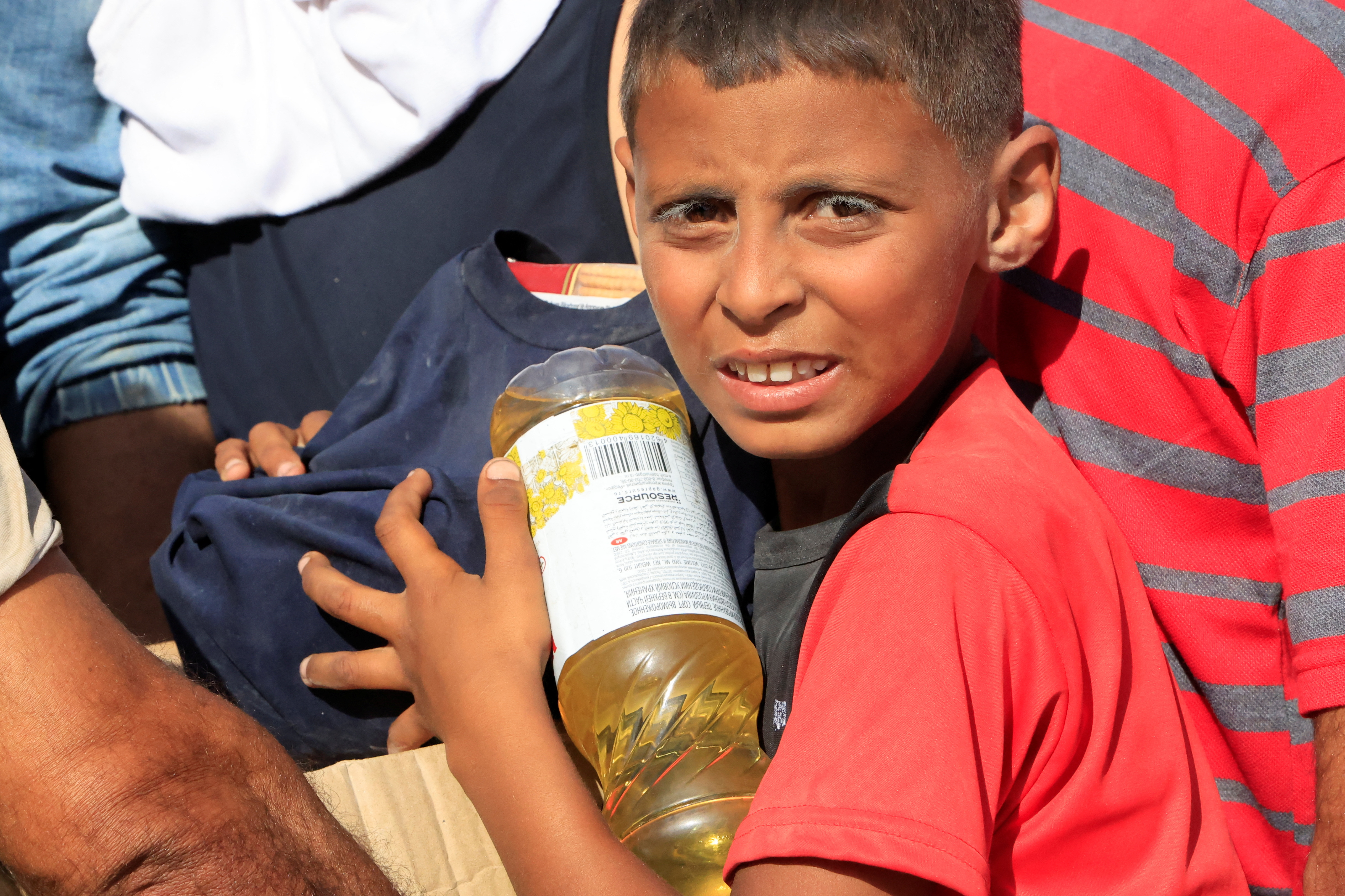 A boy cradles a clear bottle of what appears to be cooking oil in a crowded scene.