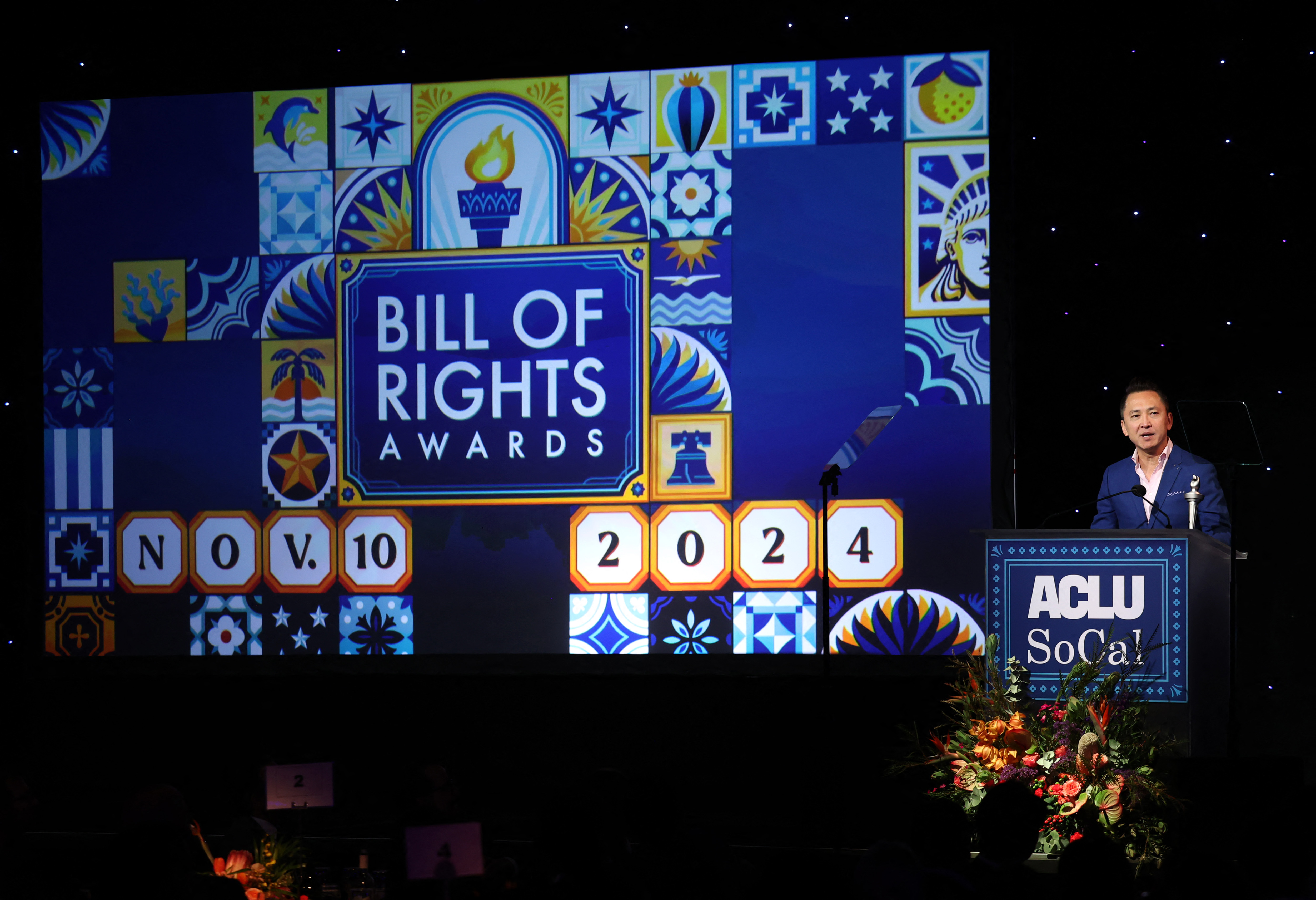 Viet Thanh Nguyen speaks on stage at the ACLU Bill of Rights Awards. A large display screen shows the name of the awards, and Nguyen stands behind a podium labelled "ACLU SoCal."