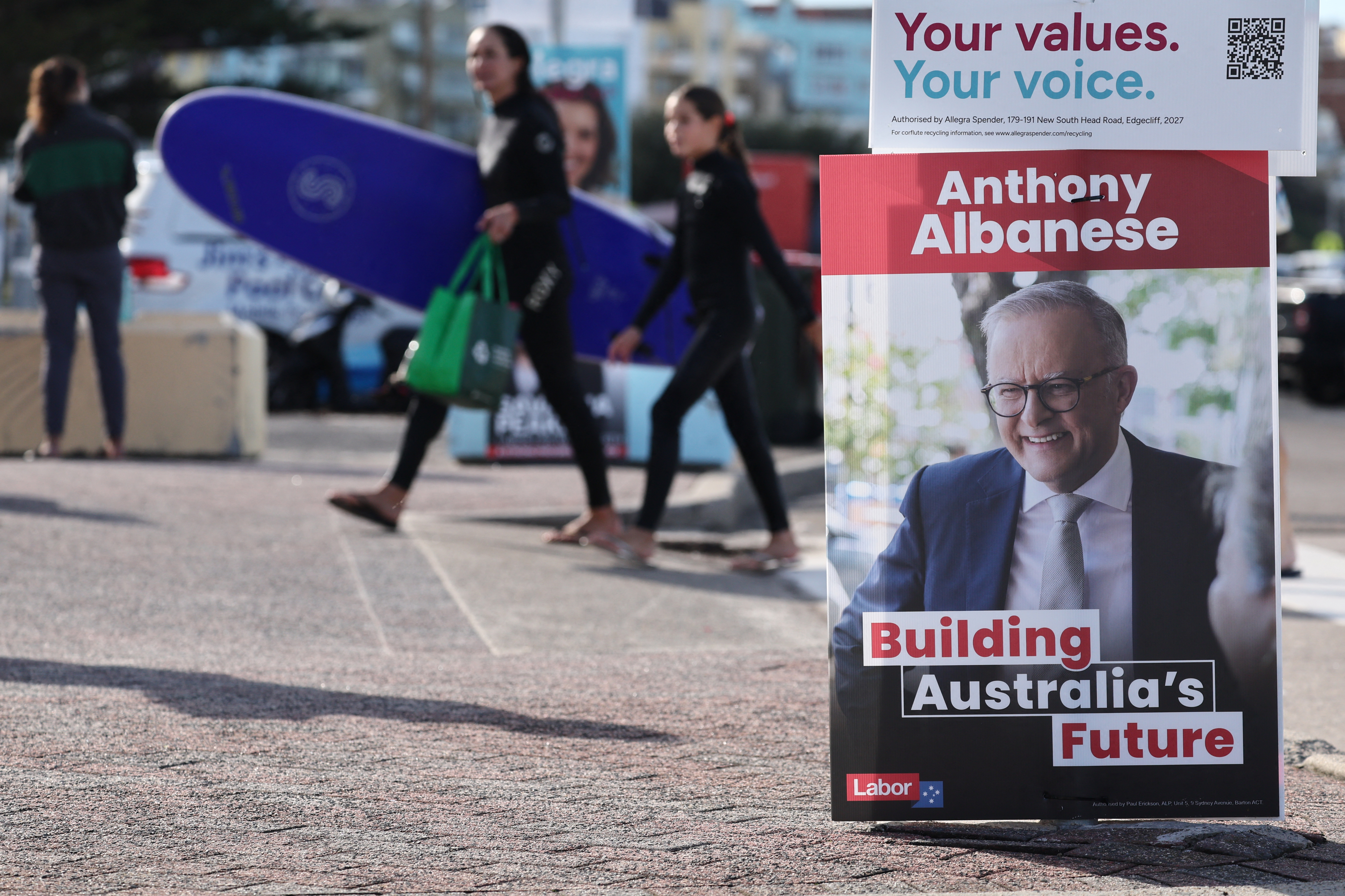A surfer carrying a surfboard passes a poster for Australia's Prime Minister Anthony Albanese on Bondi beach in Sydney on May 3, 2025. Australians started voting on May 3 in a bitterly contested general election, deciding a contest shaped by living costs, climate anxiety and Trump tariffs. (Photo by DAVID GRAY / AFP)