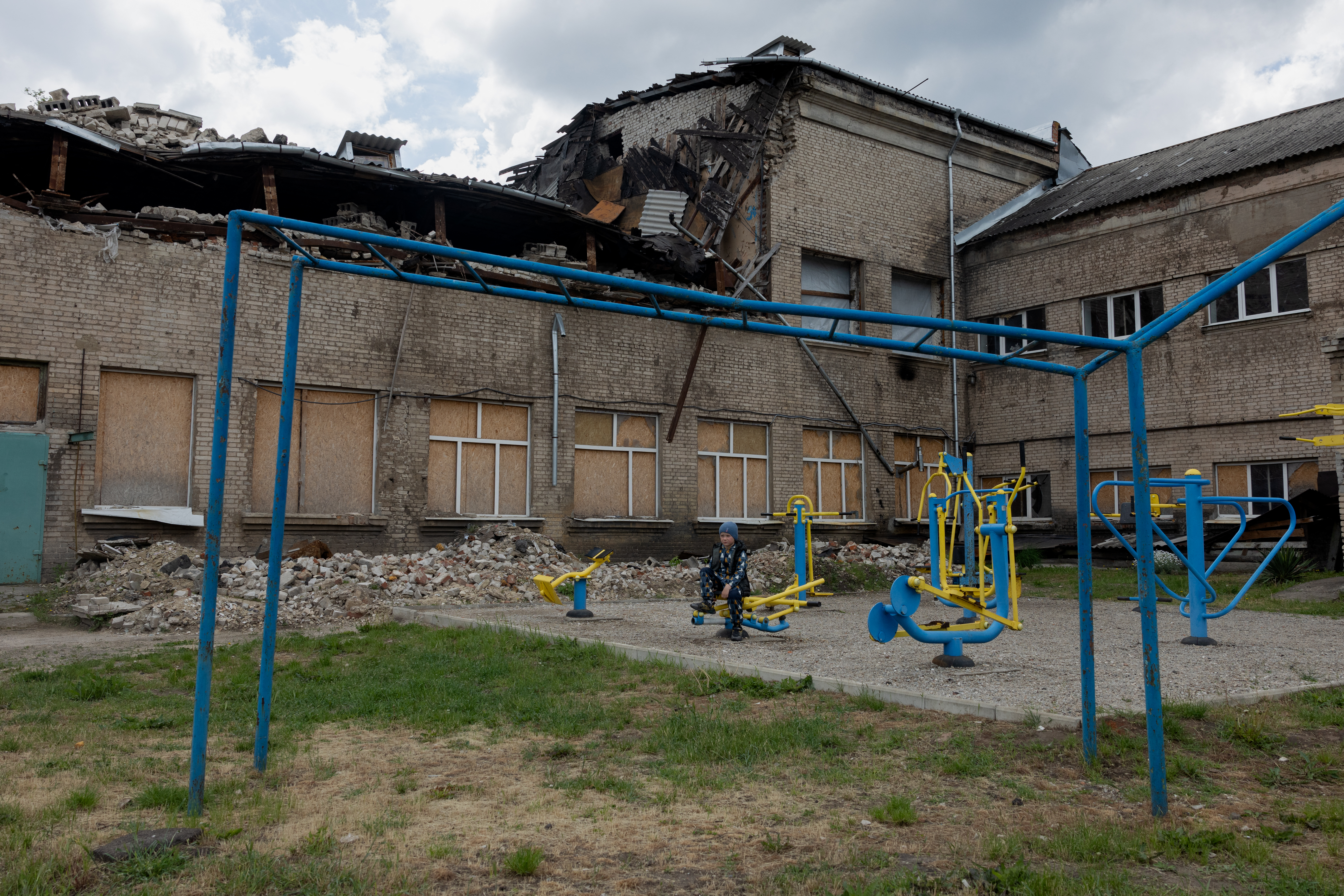 A child sits at a playground near a damaged school.