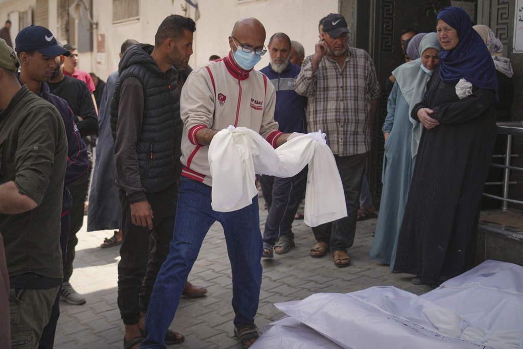 a man carries a tiny body draped in white cloth next to bodies wrapped in plastic on the ground