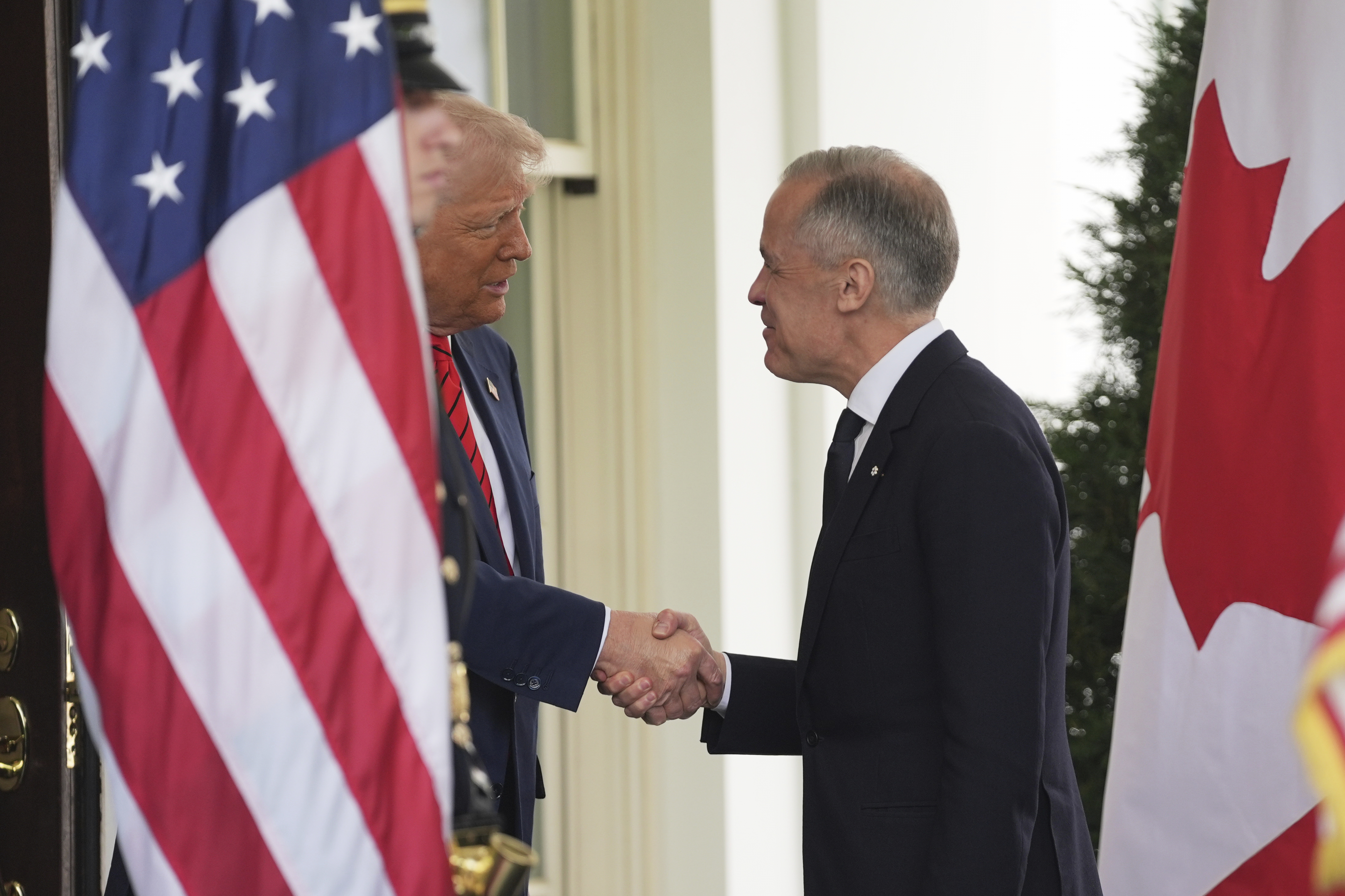 Donald Trump and Mark Carney shake hands outside the White House between a US and Canadian flag.