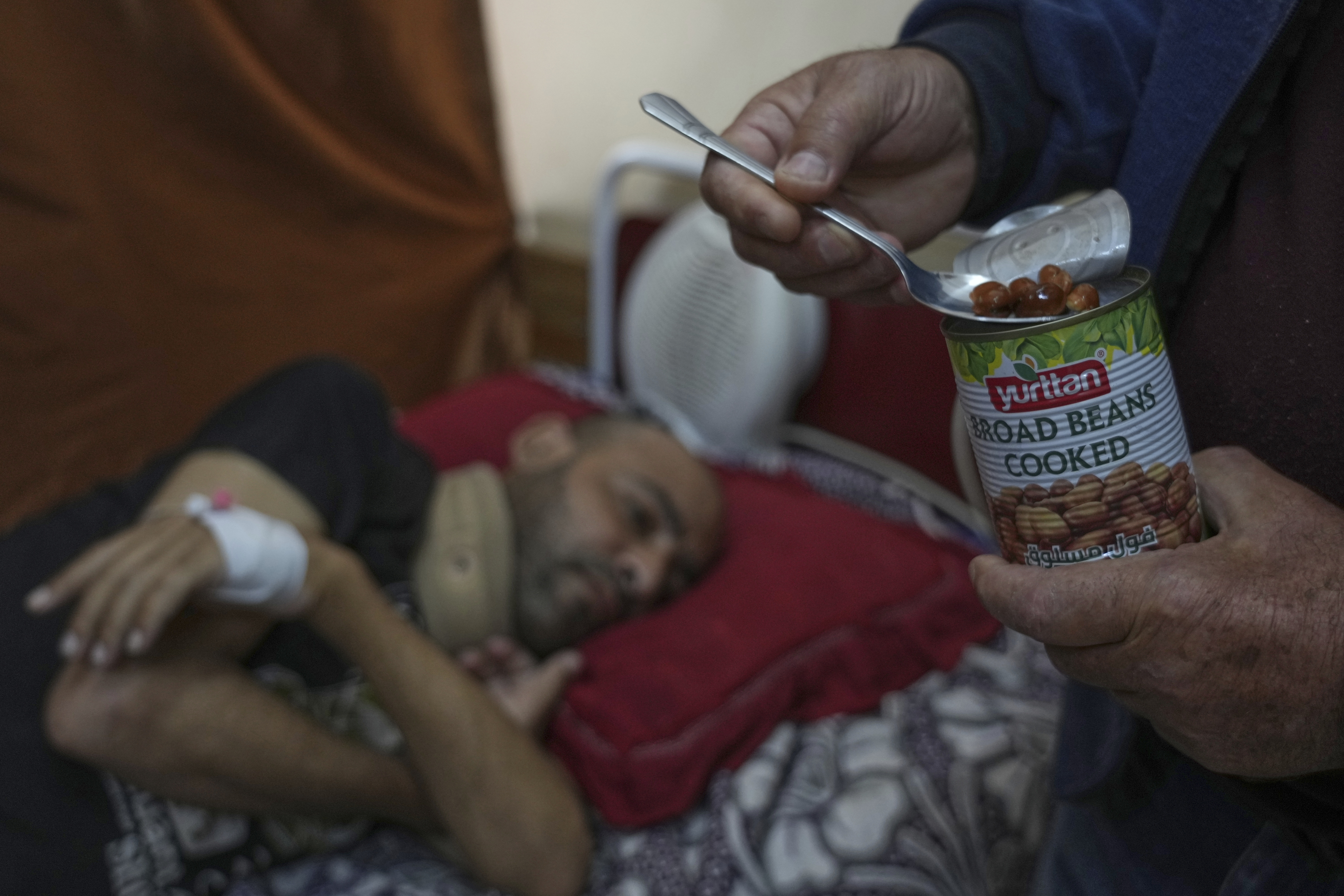 A close-up of a man's hand scooping beans from a can, while behind him, slightly blurred, lies another man on a bed, wearing a neck brace.