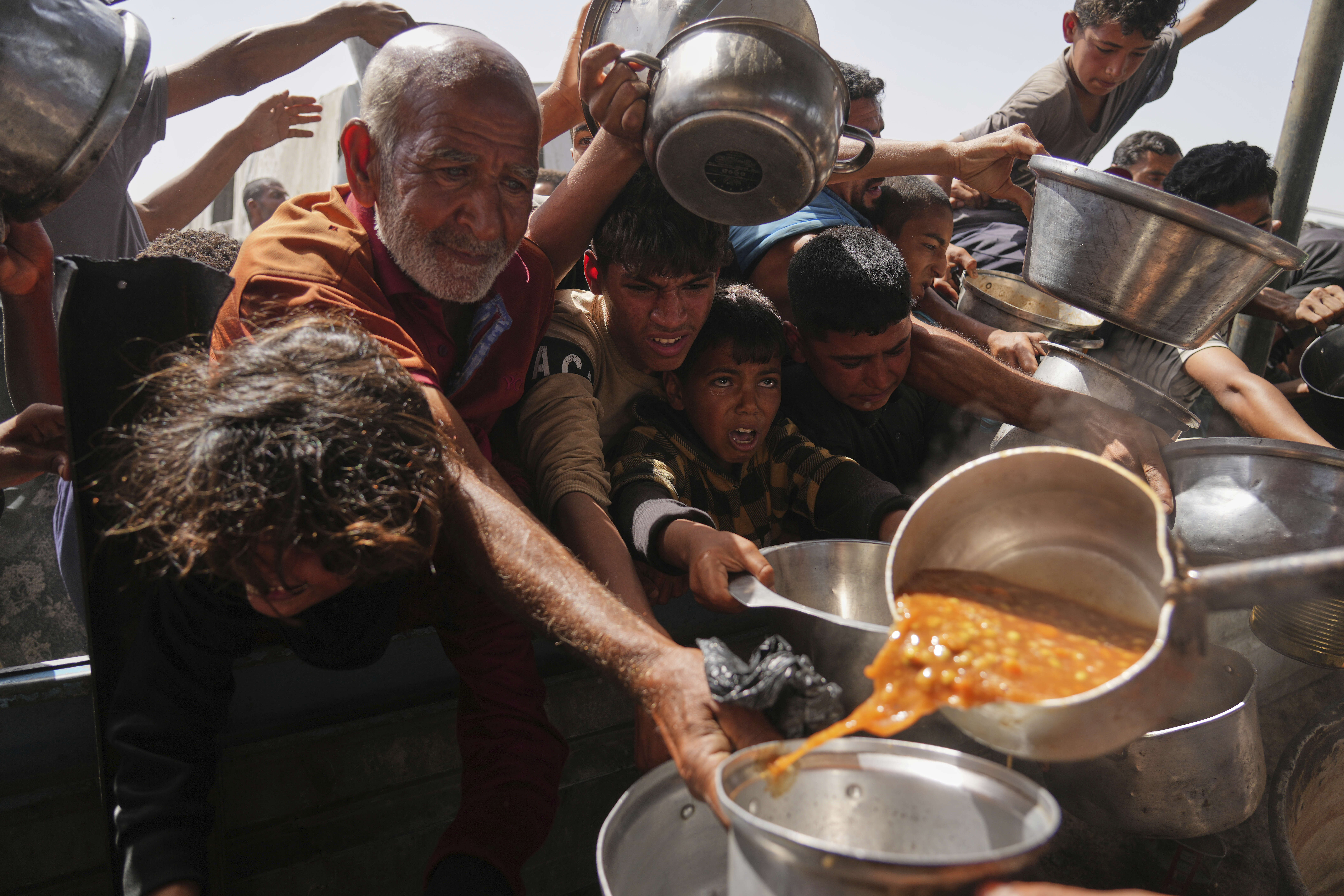 Boys and an elderly man are all holding out their pans as food is poured into them.