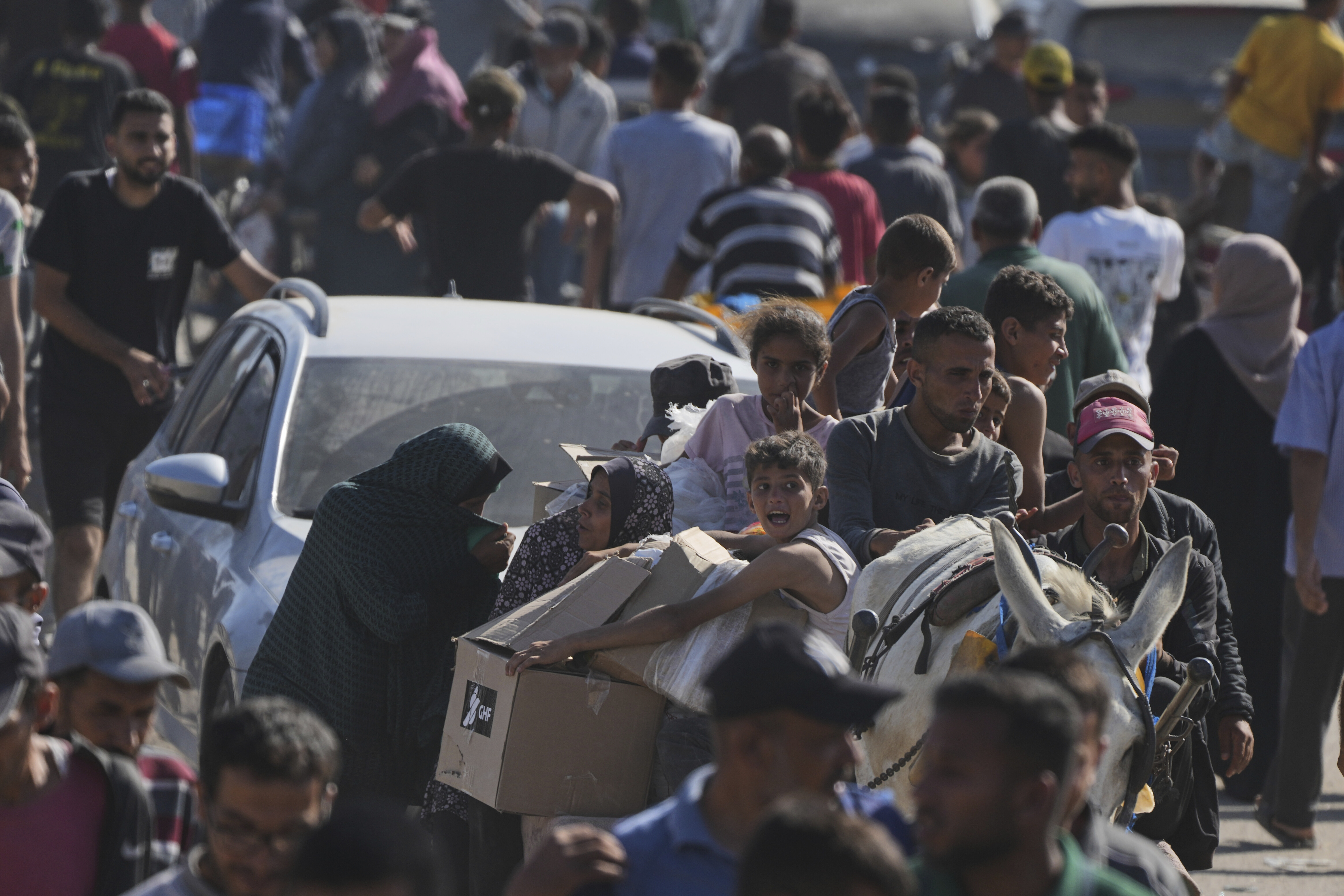 A view of the crowd receiving aid in Rafah, including a white car, a mule, a boy carrying cardboard boxes labeled GHF and many people.