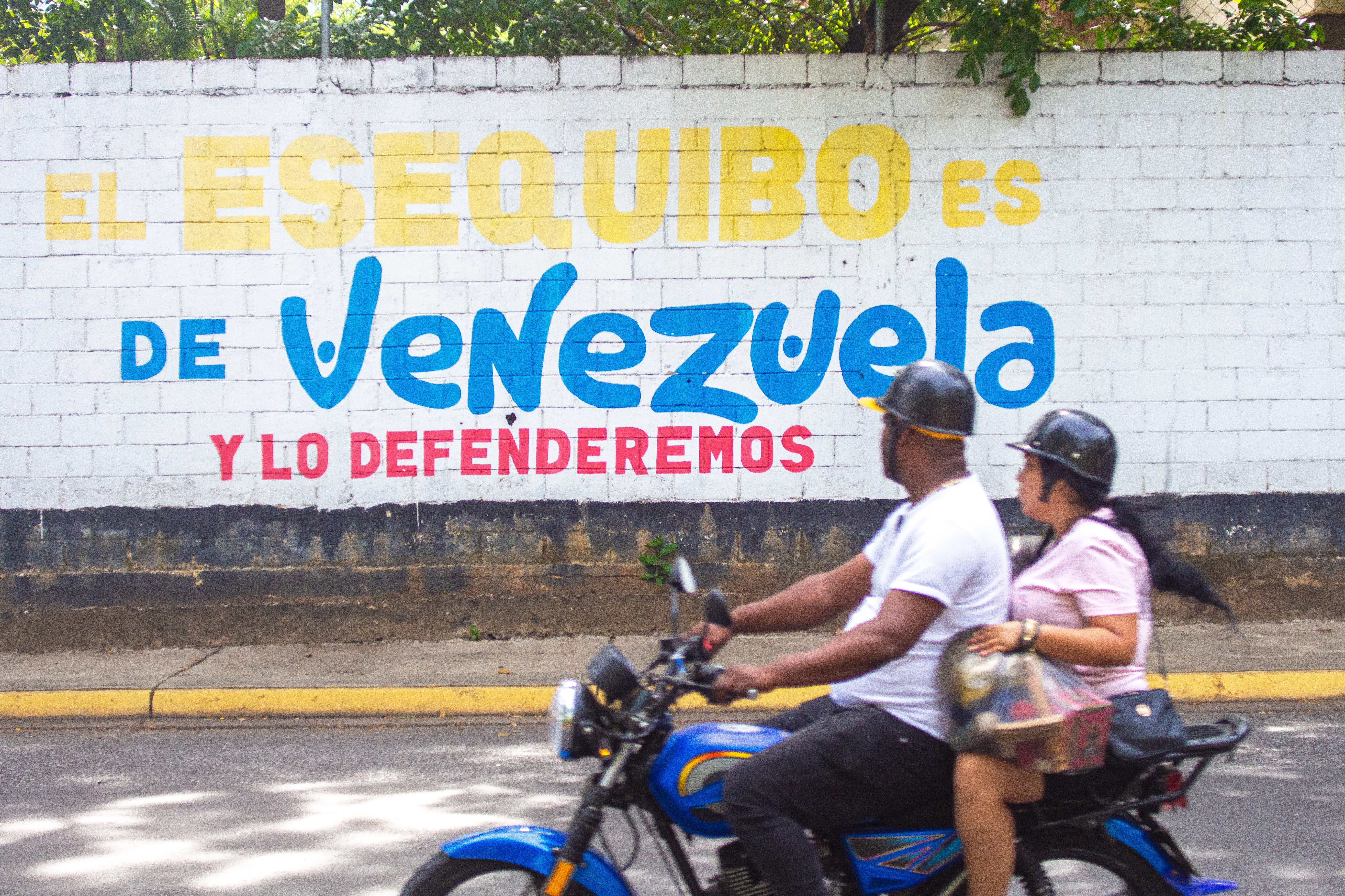 Two people on a motorcycle drive by a slogan mural
