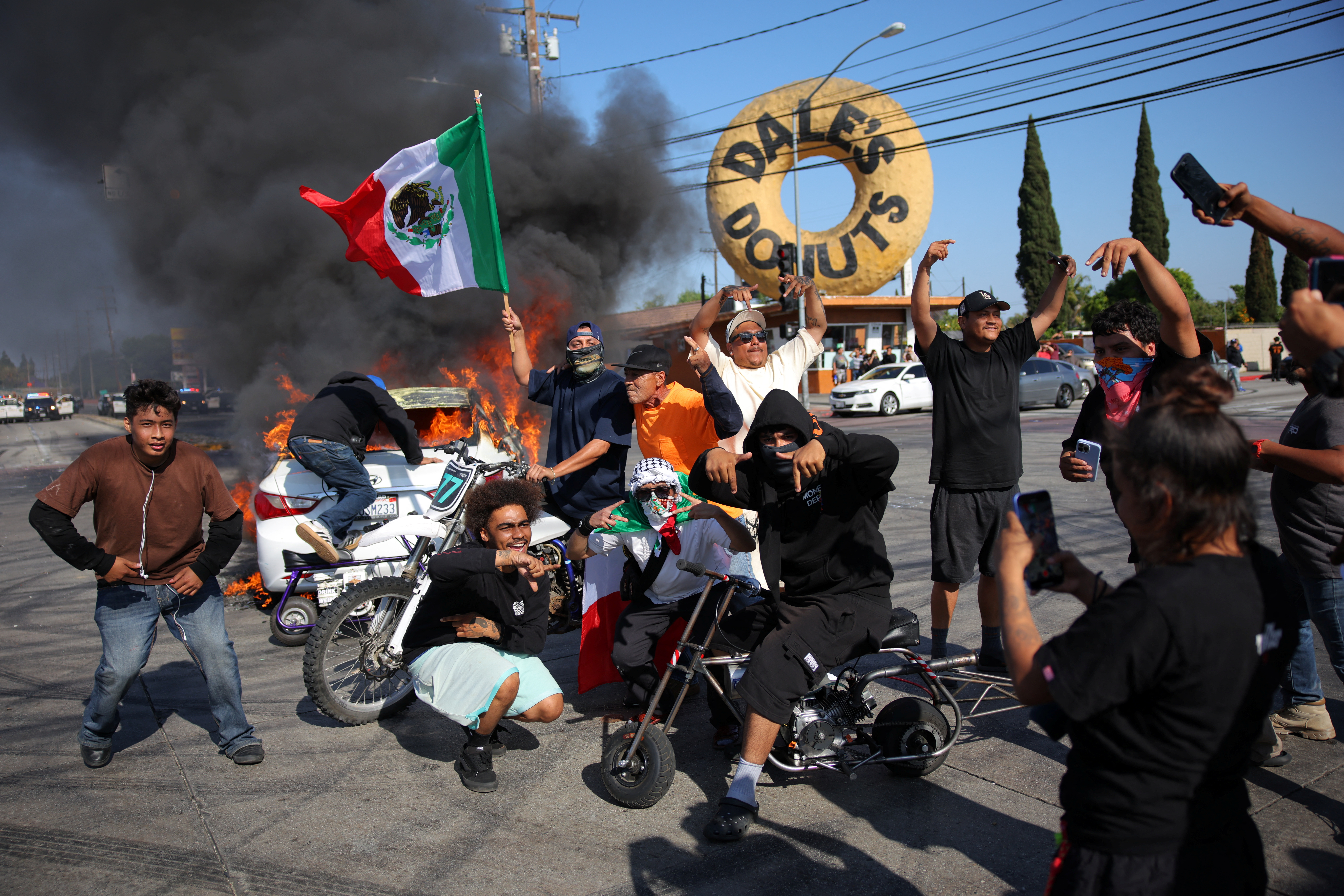 People hold Mexican flags and gesture next to a car in flames following multiple detentions by Immigration and Customs Enforcement (ICE), in the Los Angeles County city of Paramount, California, U.S., June 7, 2025.