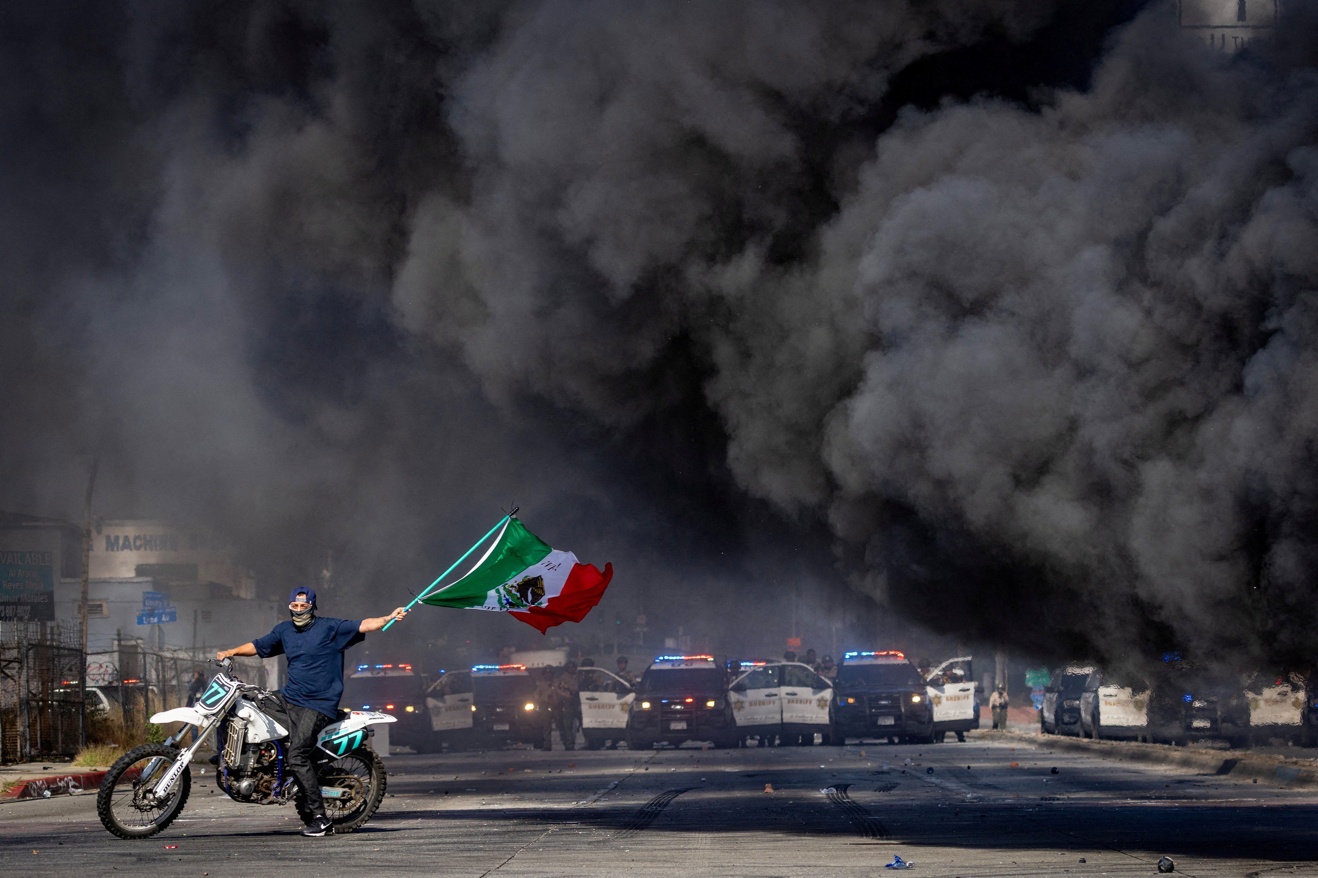 A man on a motorcycle waves a Mexican flag as smoke rises from a burning car on Atlantic Boulevard, during a standoff by protesters and law enforcement, following multiple detentions by Immigration and Customs Enforcement (ICE), in the Los Angeles County city of Paramount, California, U.S.