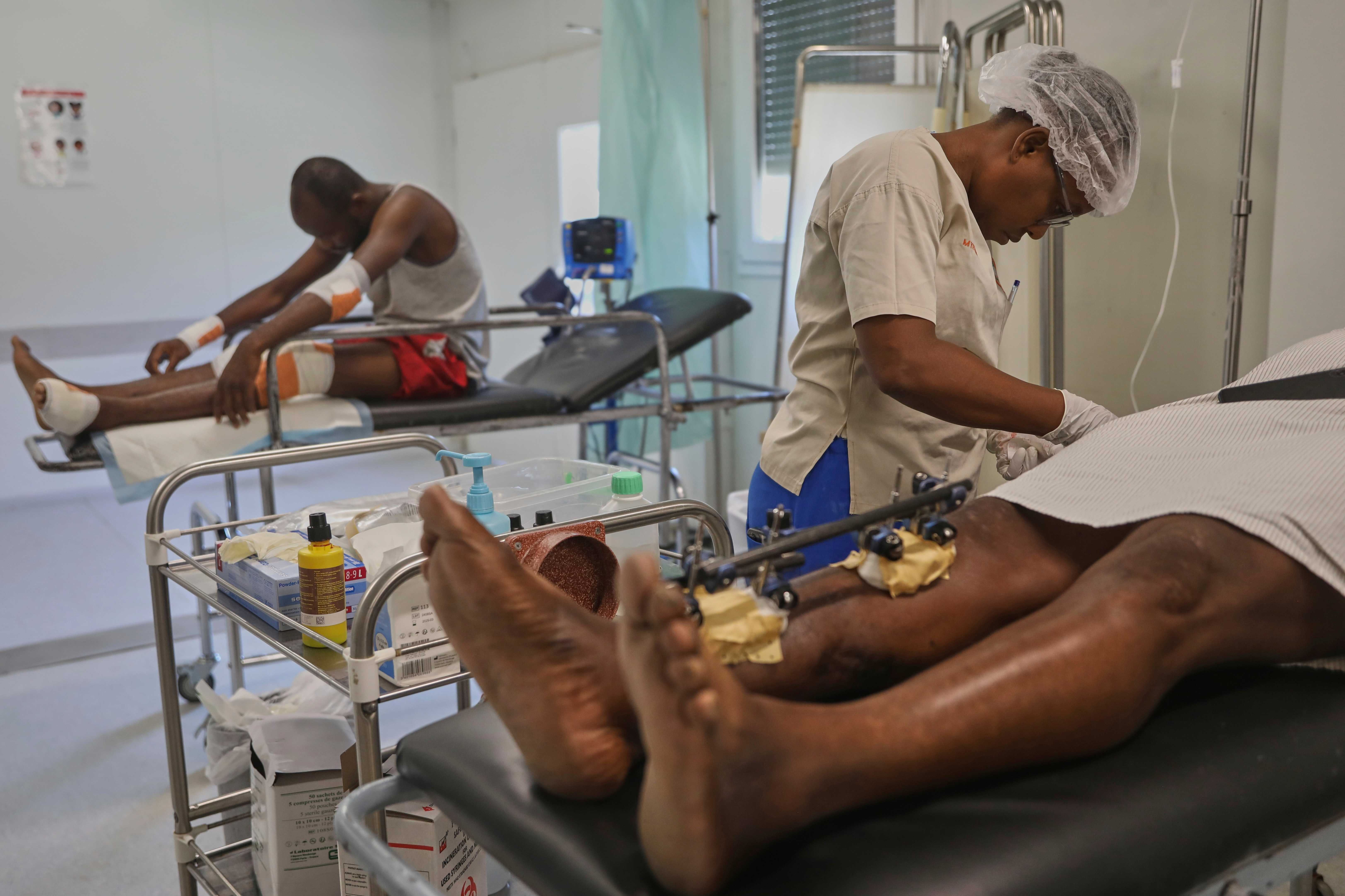 A nurse examines one patient on a hospital bed, while another on an adjacent bed leans forward.